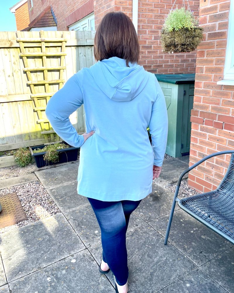 A lady standing in a garden in front of a fence with her back to the camera wearing a light blue hooded tunic top