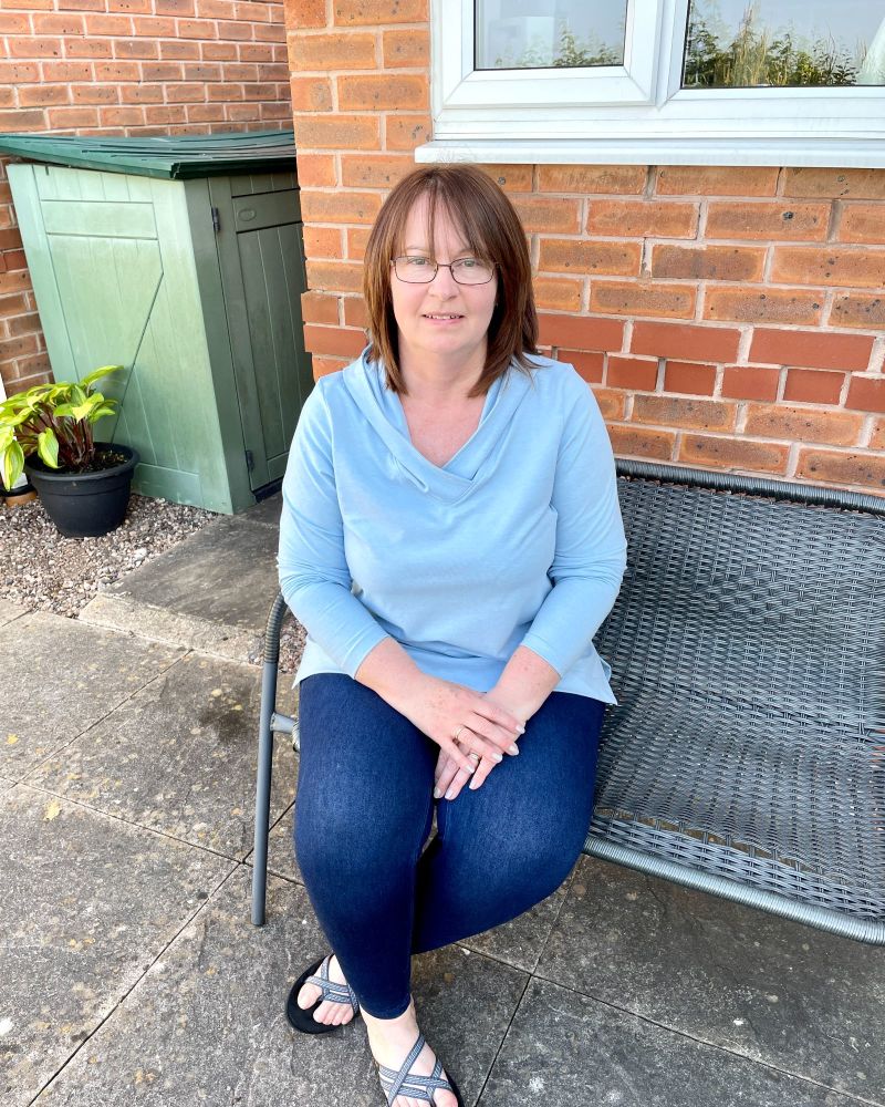 A lady sitting on a garden bench wearing a light blue tunic top and jeans