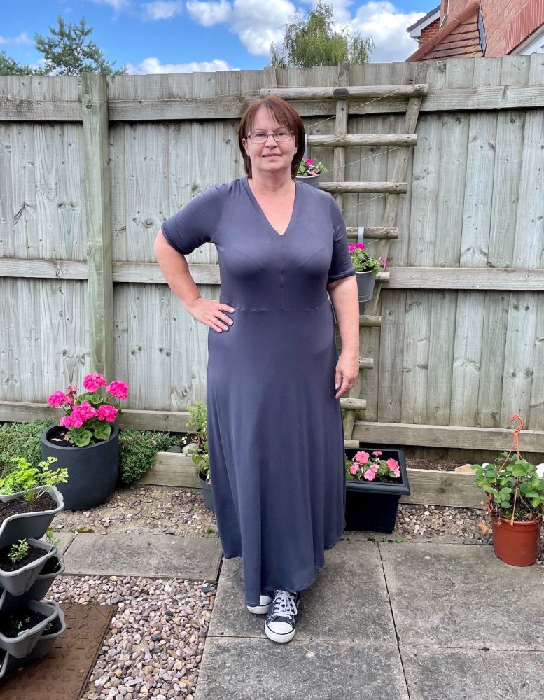 A lady wearing the maxi length Olympia Dress in blue Jersey standing in front of plants and a fence.