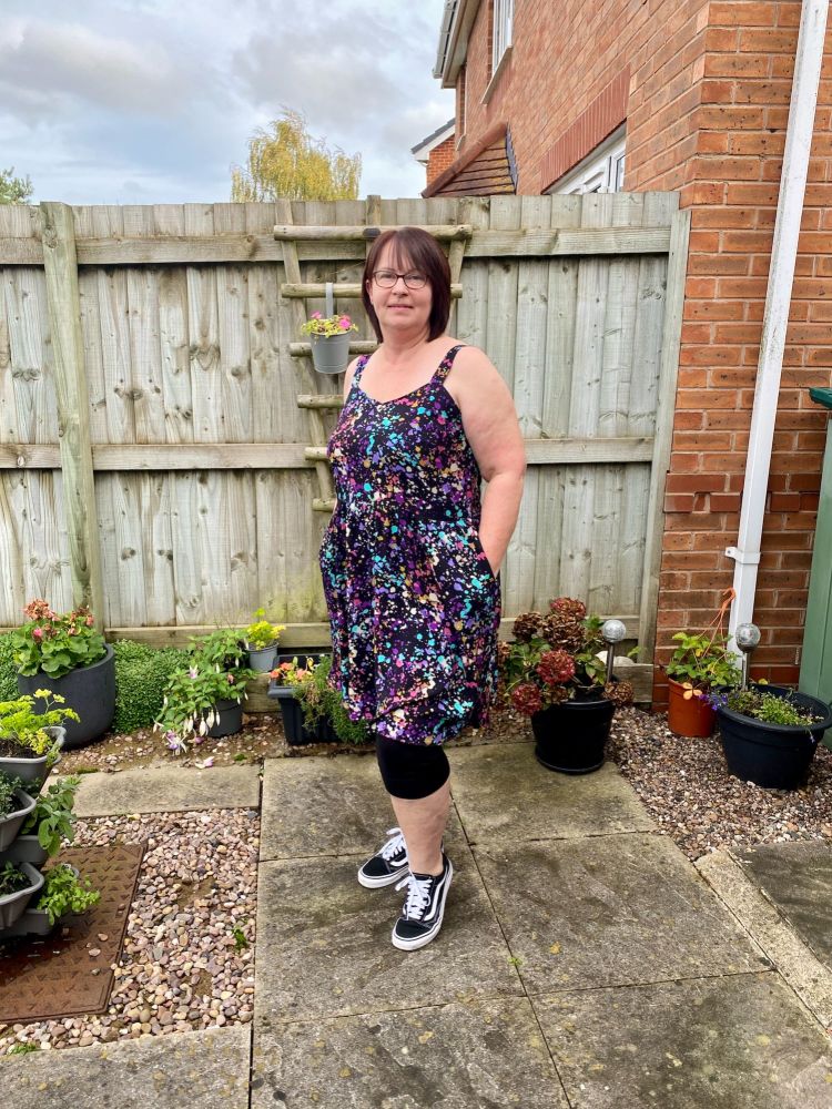 A lady with short brown hair standing in front of a fence wearing a black and multicoloured Sundress