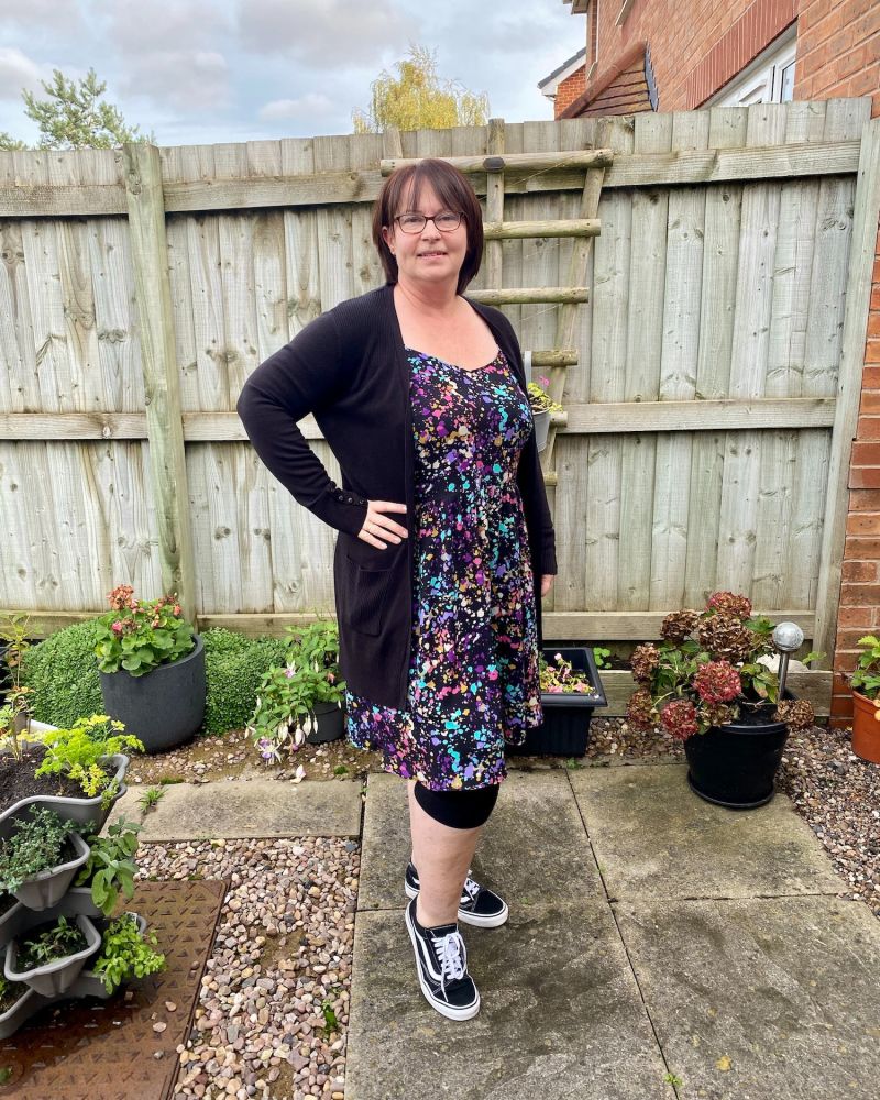 A lady with short brown hair standing in front of a fence wearing a black and multicoloured Sundress & black cardigan, leggings and pumps