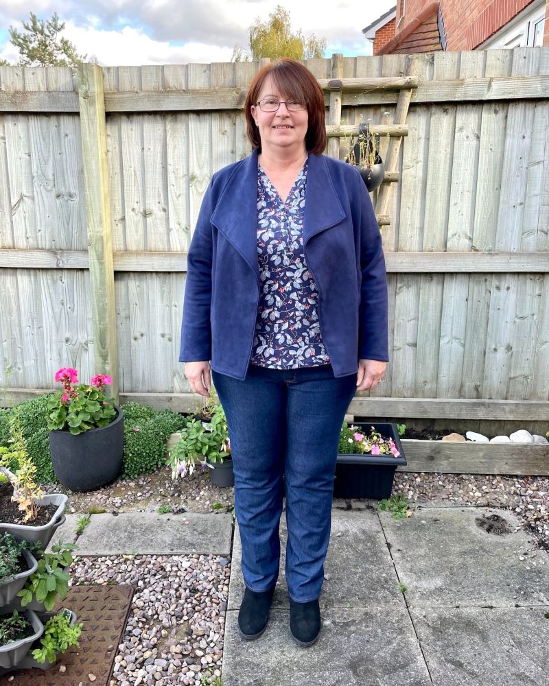 A lady with short brown hair is standing in front of a garden fence wearing a pair of blue Legato Jeans and a blue Jacket