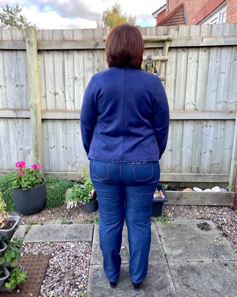 A lady with short brown hair standing in front of a garden fence wearing a pair of dark blue jeans and a navy jacket showing the back view