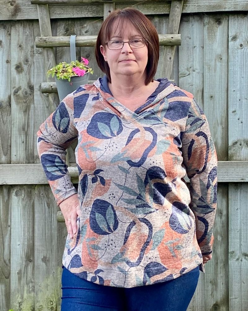 A lady standing in a garden in front of a fence wearing a patterned tunic top & jeans