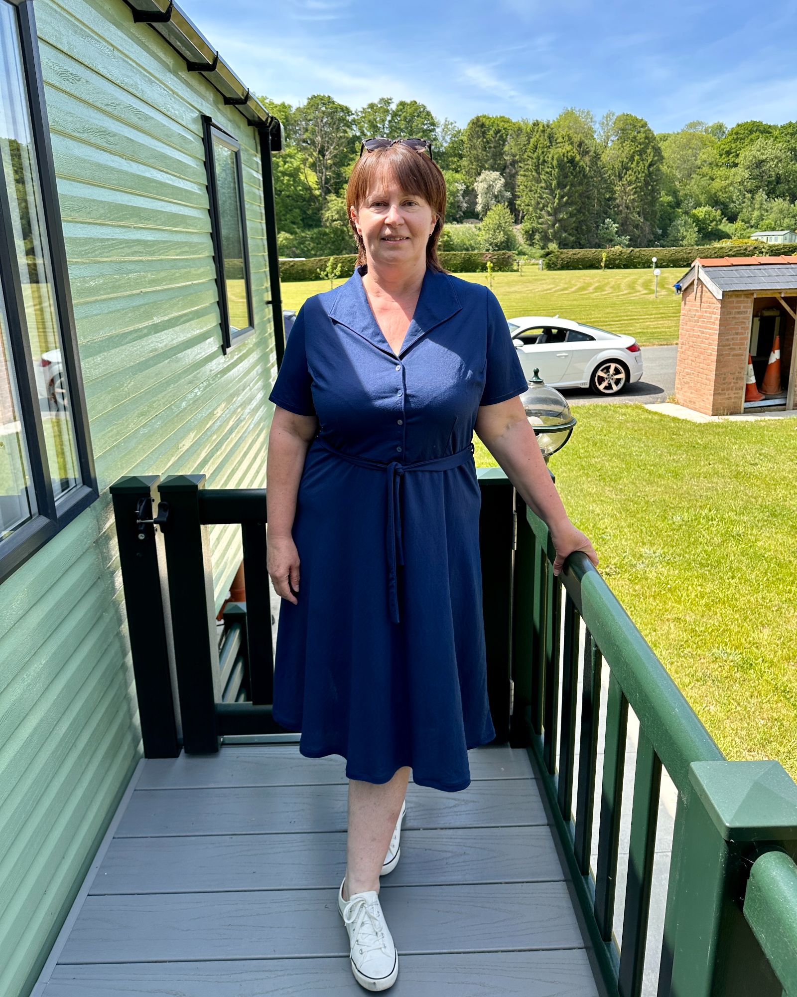A lady with short brown hair standing on a caravan decking wearing a navy jersey dress. & white pumps.