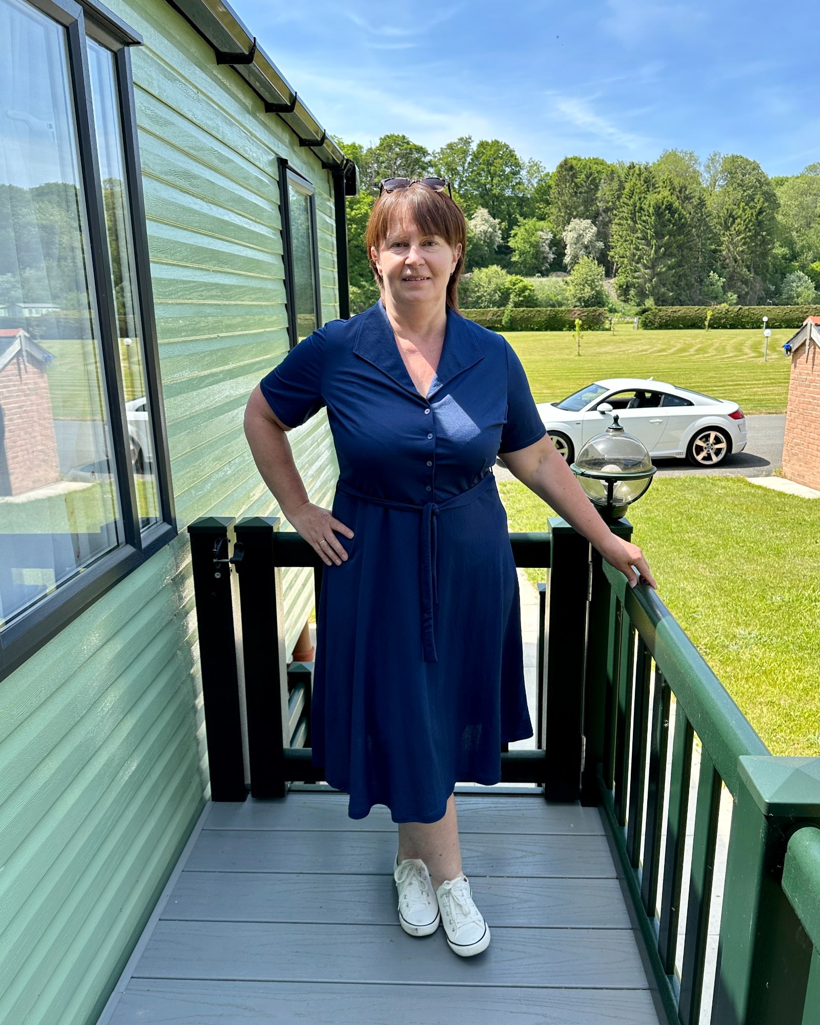 A lady with short brown hair standing on a caravan decking wearing a navy jersey dress.