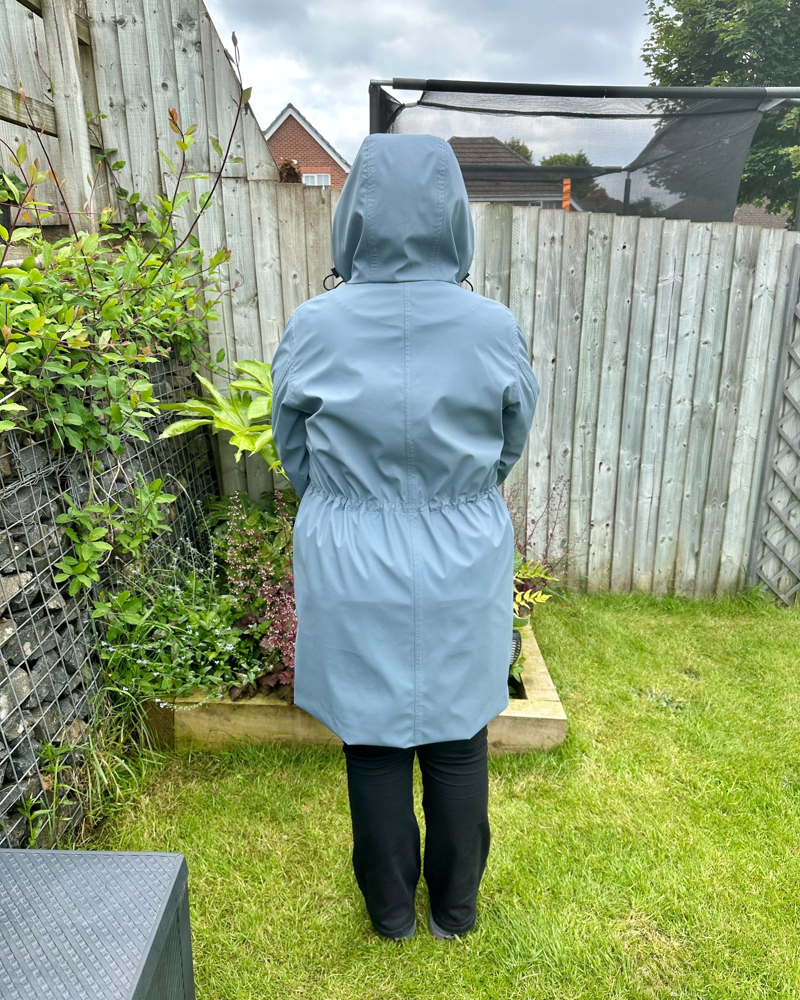 A lady standing in a garden with her back to the camera wearing a blue grey raincoat with the hood up