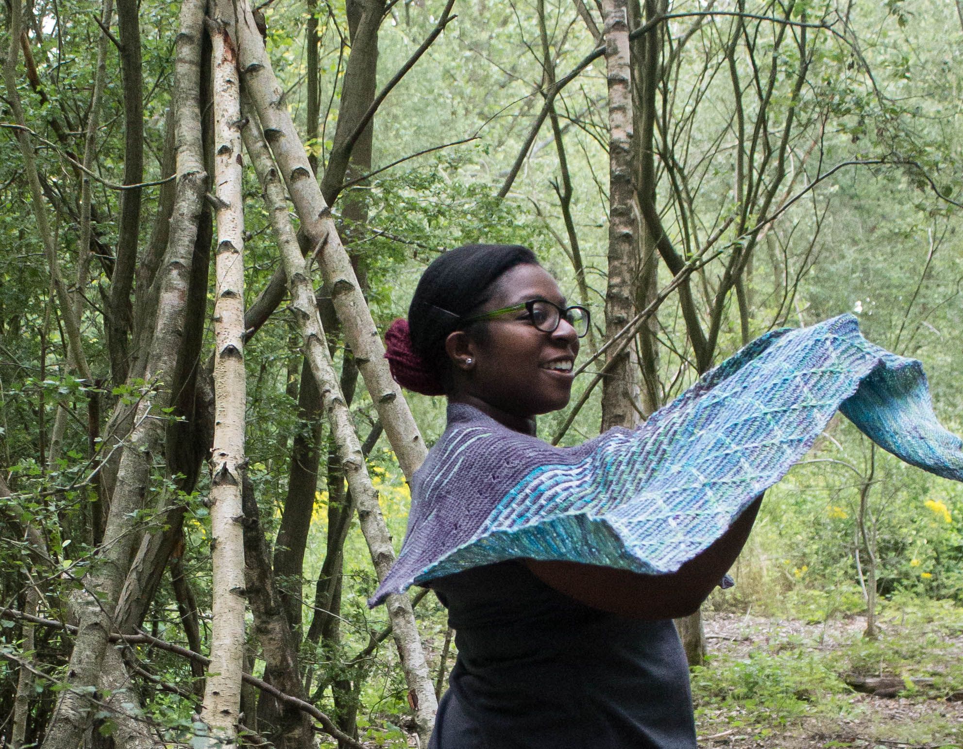 Image of a black woman smiling, holding the edge of a shawl as it floats in the wind, with the rest wrapped around her. Showing off the slipped stitch patterning and cables.