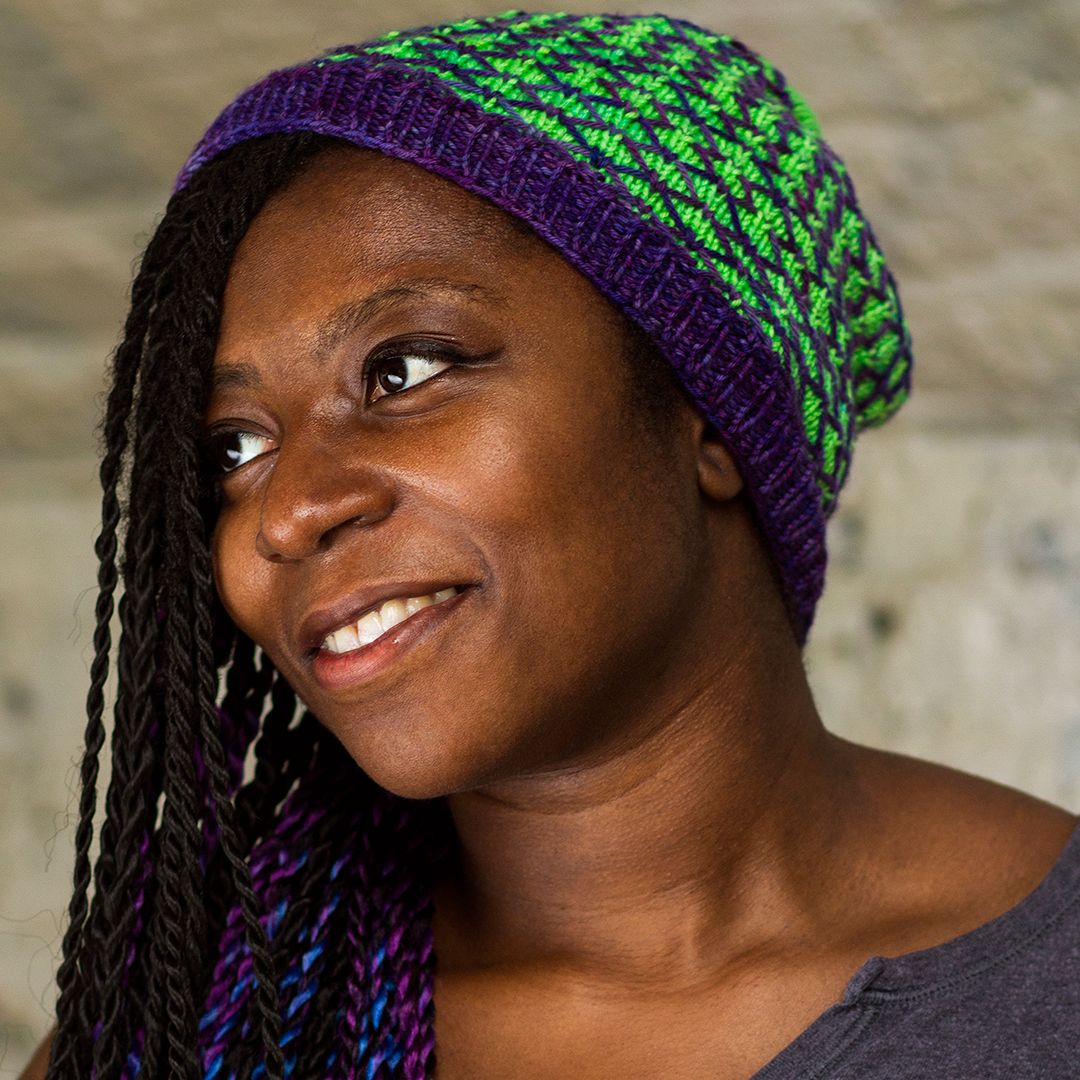 Picture of lola, a black person wearing a green and purple hat smiling looking to the right side of the picure