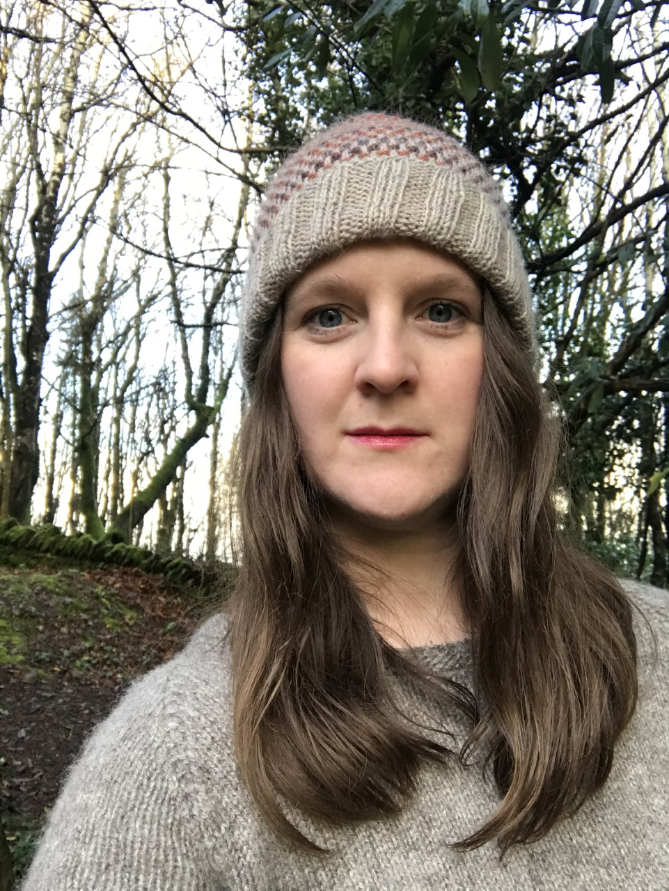 picture of helena, a brown haired white woman, staring at the camera,wearing a woolly hat, trees in the background