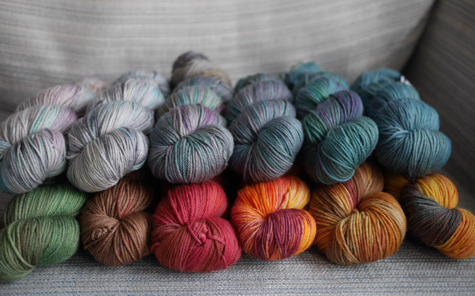  On a grey and blue striped fabric background,  12 skeins of dk weight yarn sit 2 skein high in a line. with colours ranging from silver with pastel flashes to a deep steely blue and cerulean, green and purple flashes on the top. On the bottom colours range from greens and browns to bright orangey reds with a shot of black.