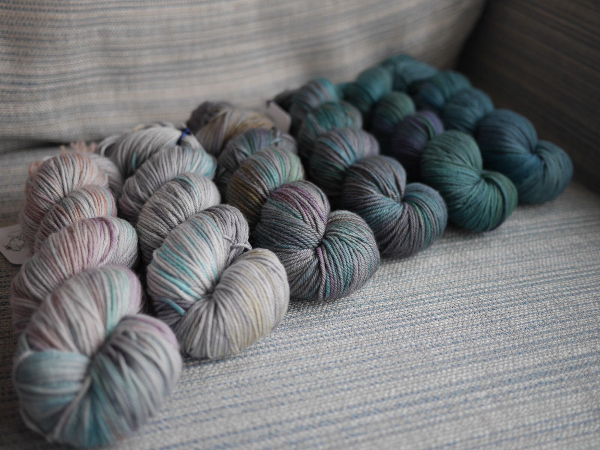 6 skeins on a grey blue striped background , at an angle colourways from left to right are the Air and Water Months in order