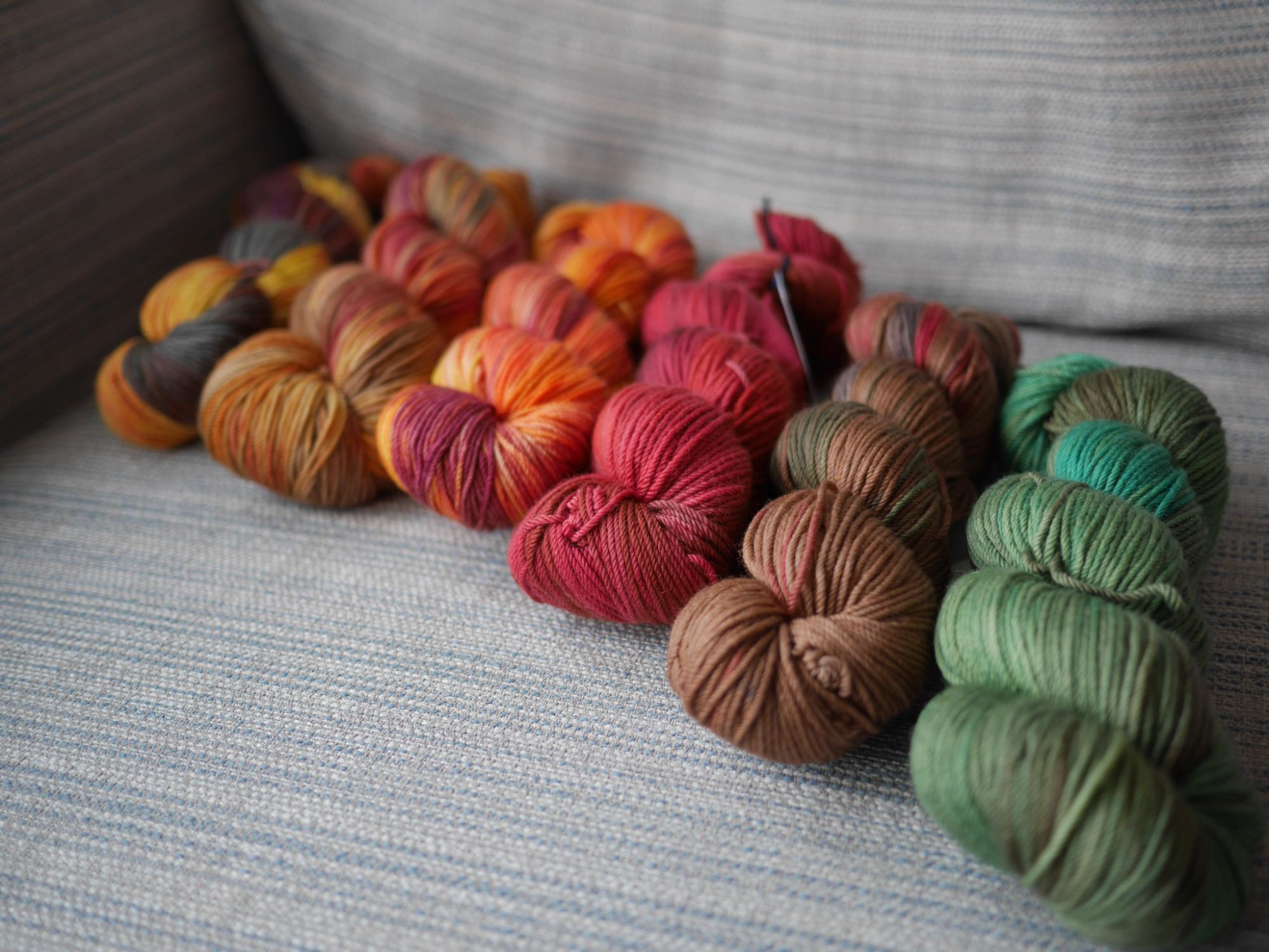 6 skeins on a grey blue striped background , at an angle colourways from right to left are the Earth and Fire Months in order