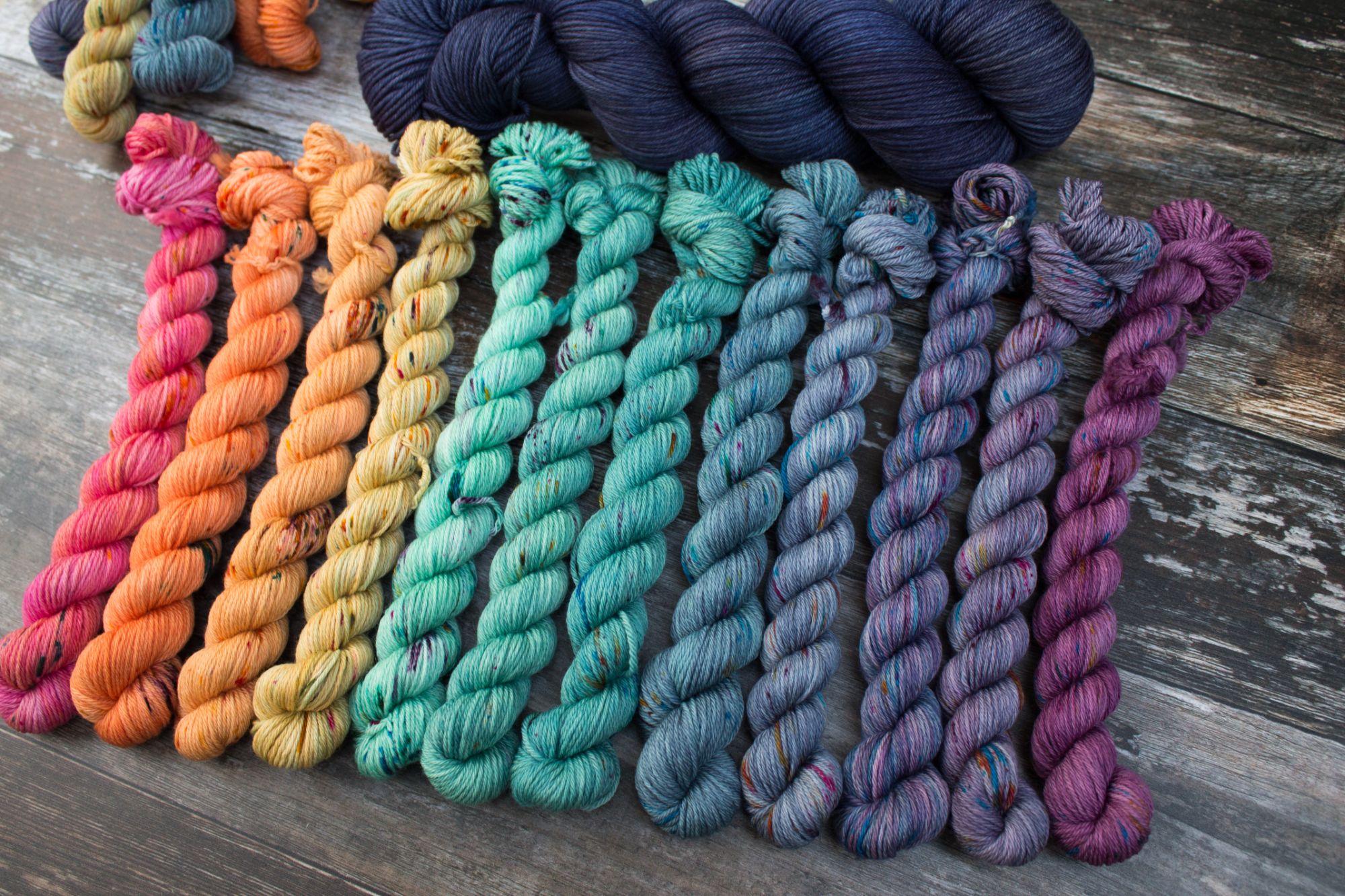 A selection of mini skeins in a gradient from orange teal to blue purple with a dark blue skein above them