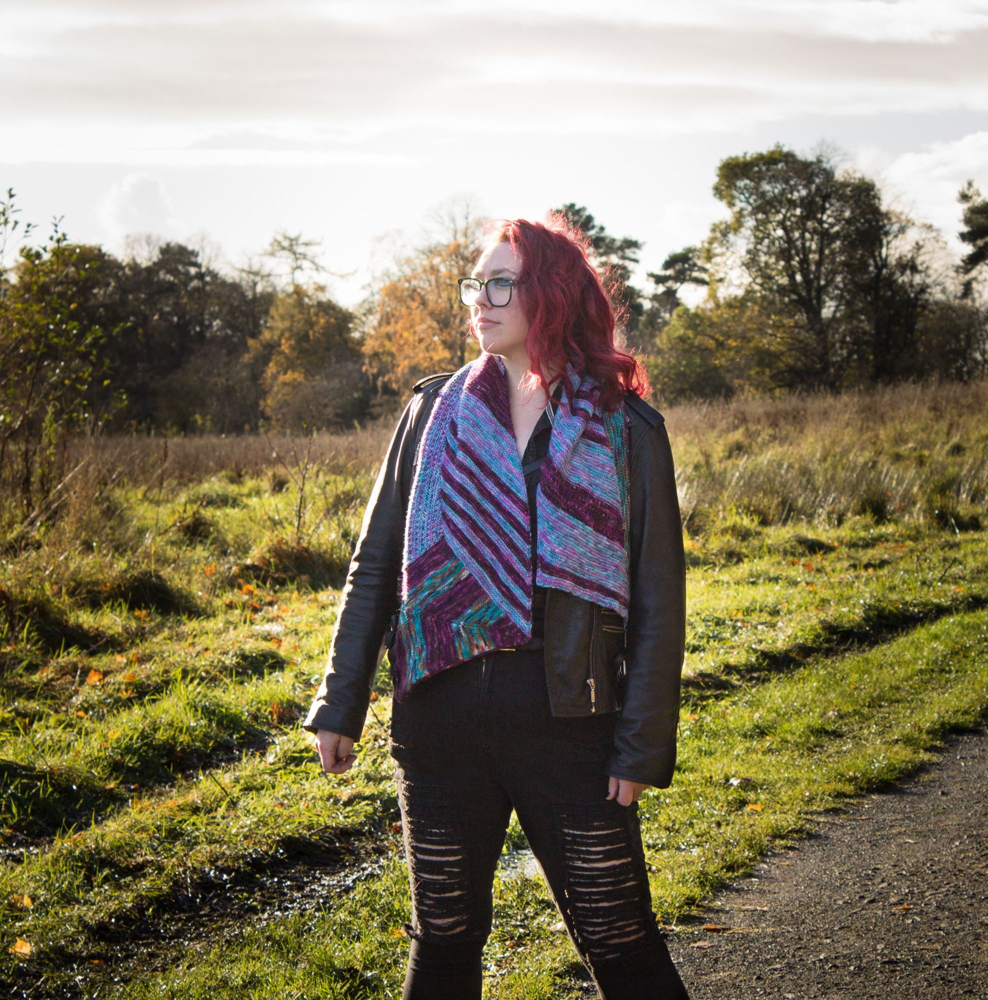 Ryann (red haired white woman), standing boldy in the centre of a grassland path, wearing the Meteor shawl draped around her neck.