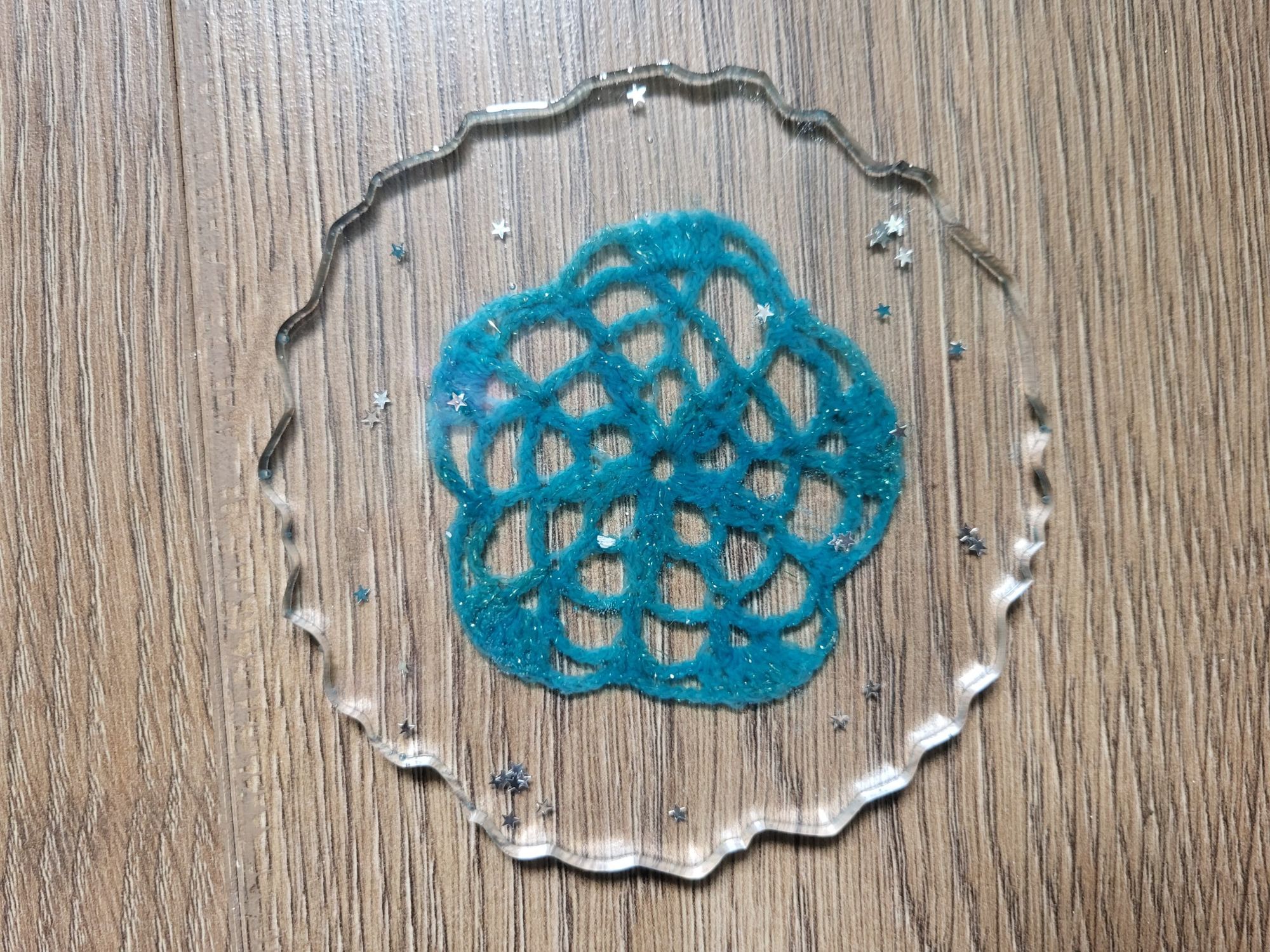 a clear resin coaster sits in the middle of the image with a blue crocheted snoflake captured in it with tiny silver snowflakes also scattered inside the resin