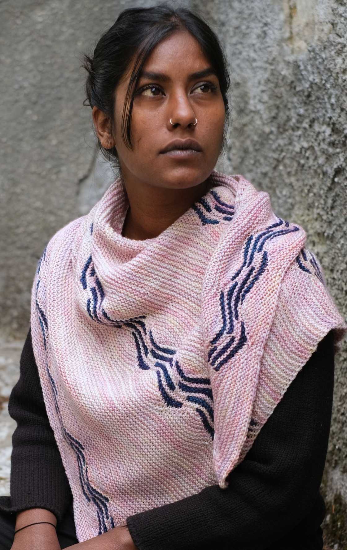 brown skinned person with hair pulled back from their face, they havce some strands in front and a a nose ring on either side of their nose. they are looking forward, up and to the left of the camera. They wear a pink knitted shawl draped around their shoulderd which features dark blue sectional waves. 