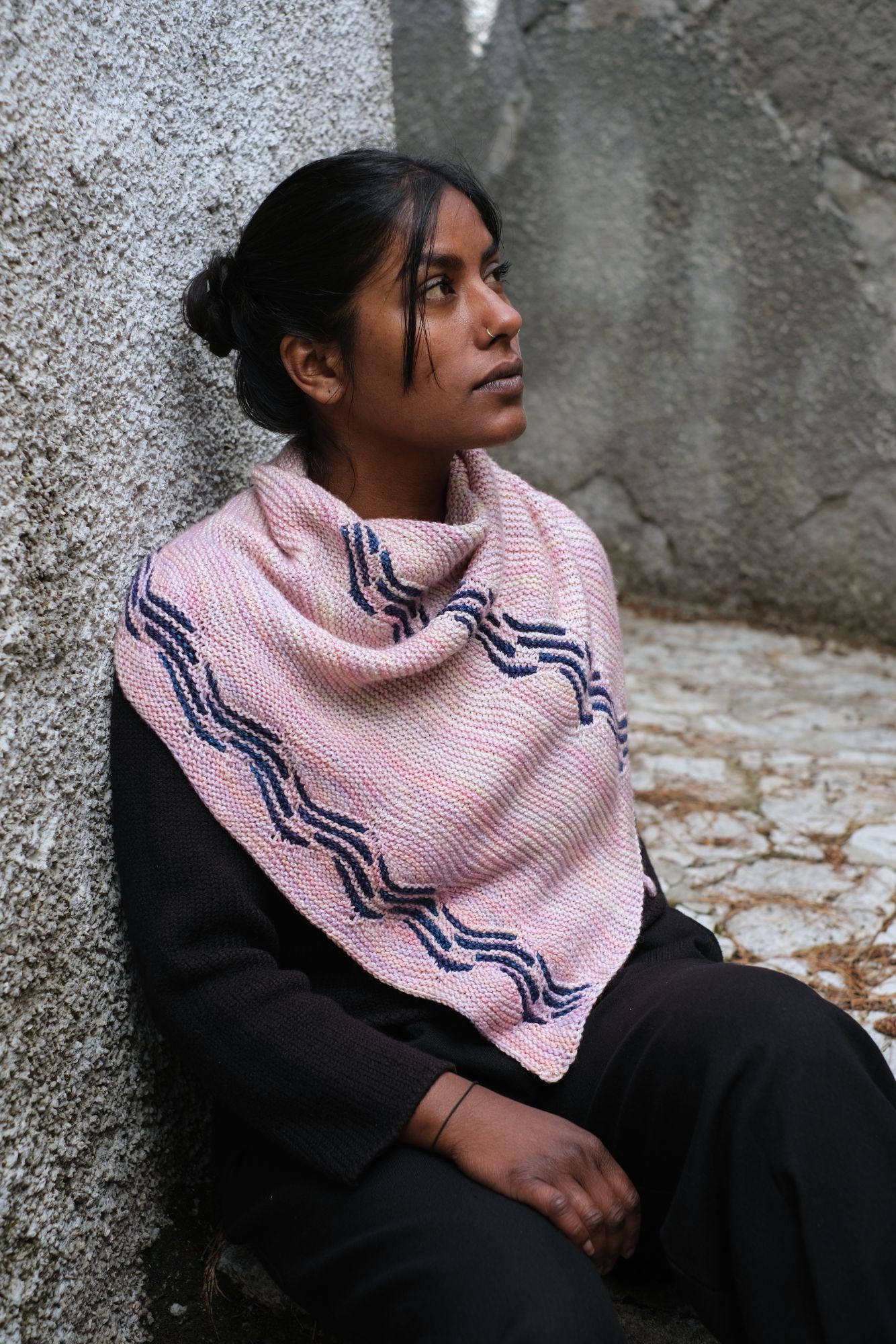 brown skinned person with hair pulled back from their face, they havce some strands in front and a a nose ring on either side of their nose. They are  facing to the side and looking up and away. They wear a pink knitted shawl draped around their shoulderd which features dark blue sectional waves. 