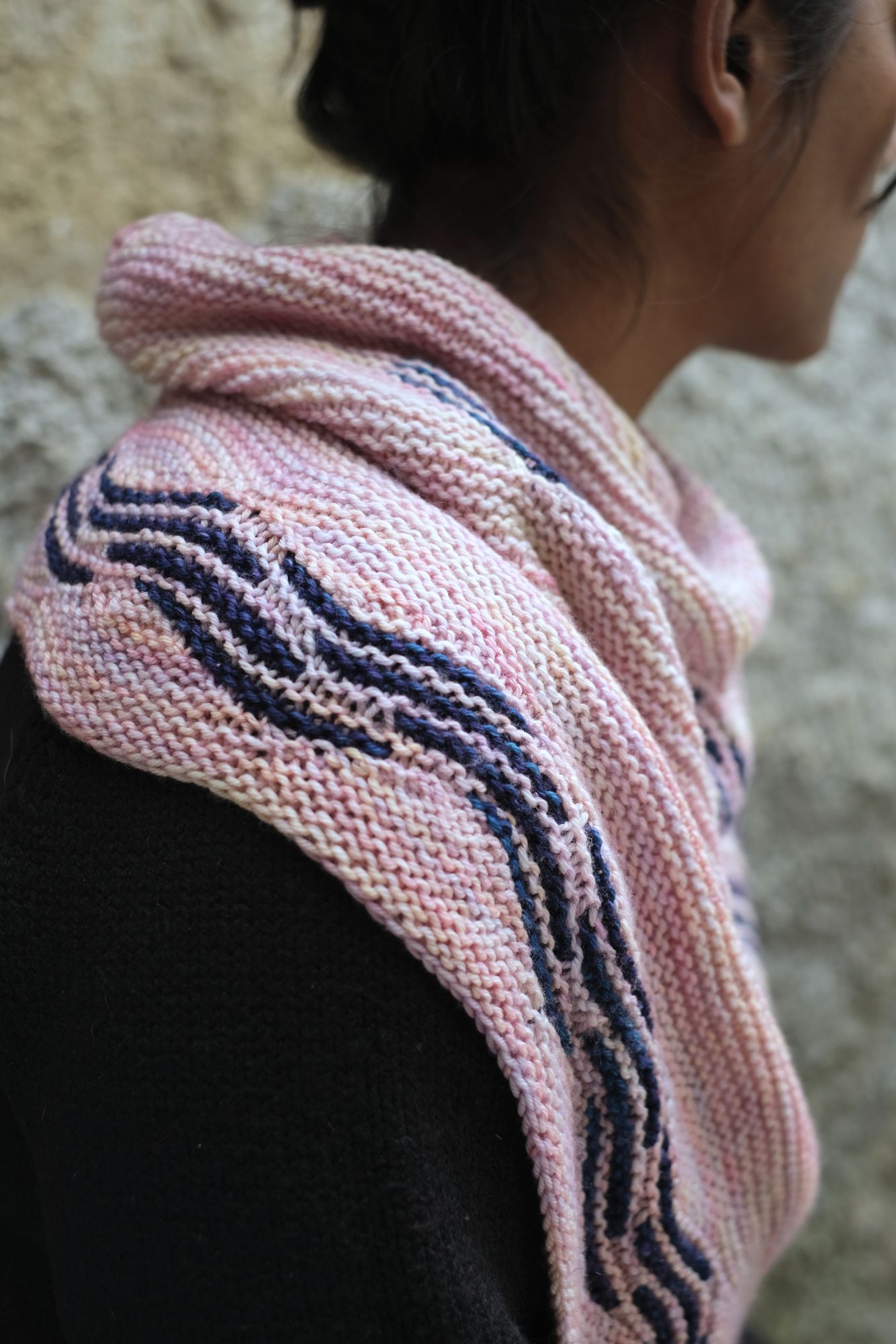  pink knitted shawl draped around their shoulderd which features dark blue sectional waves. close up of the shawl