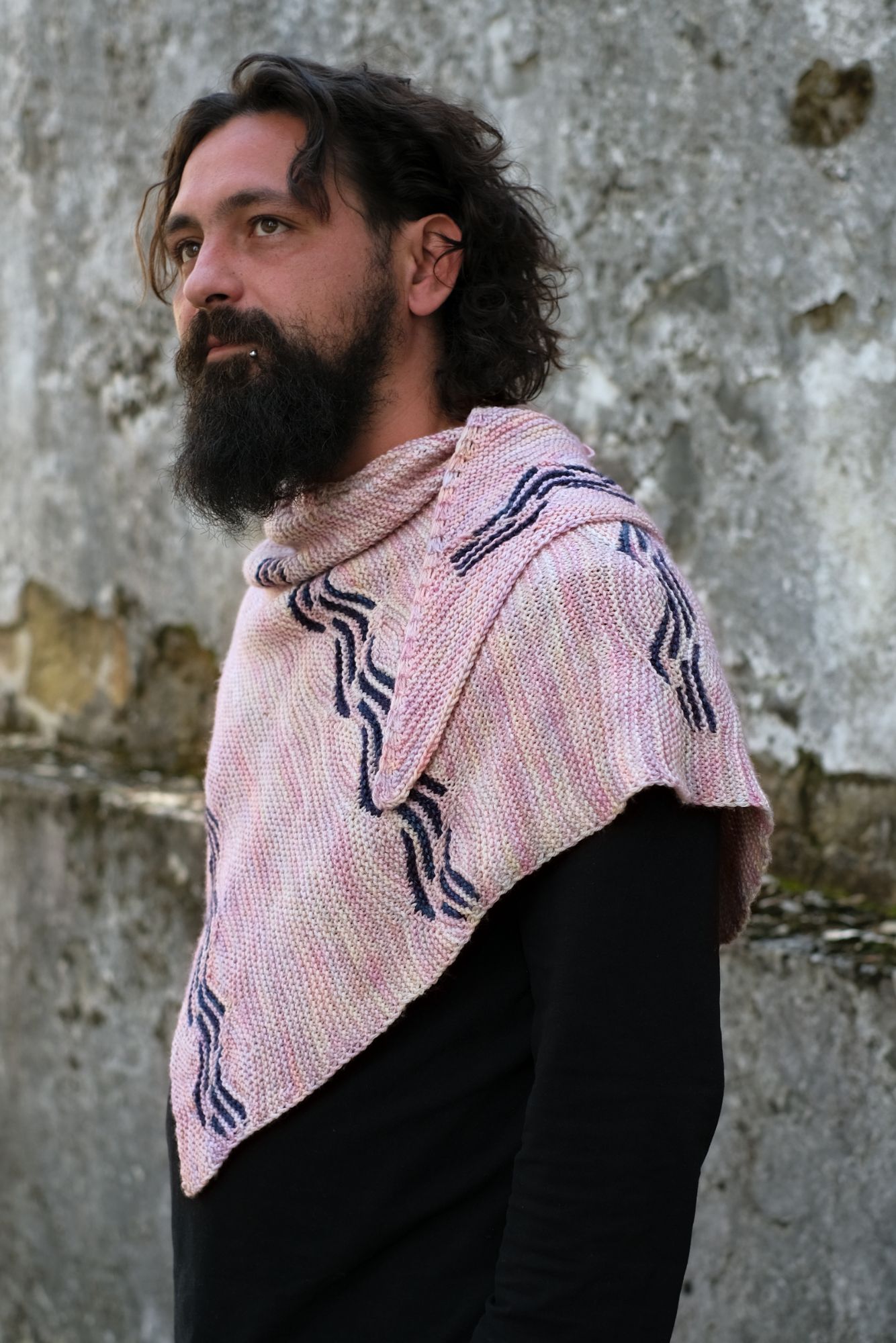 white person with a beard and lip ring, faces to the side and out wearing a pink shawk over their shoulders with blue short row motifs 
