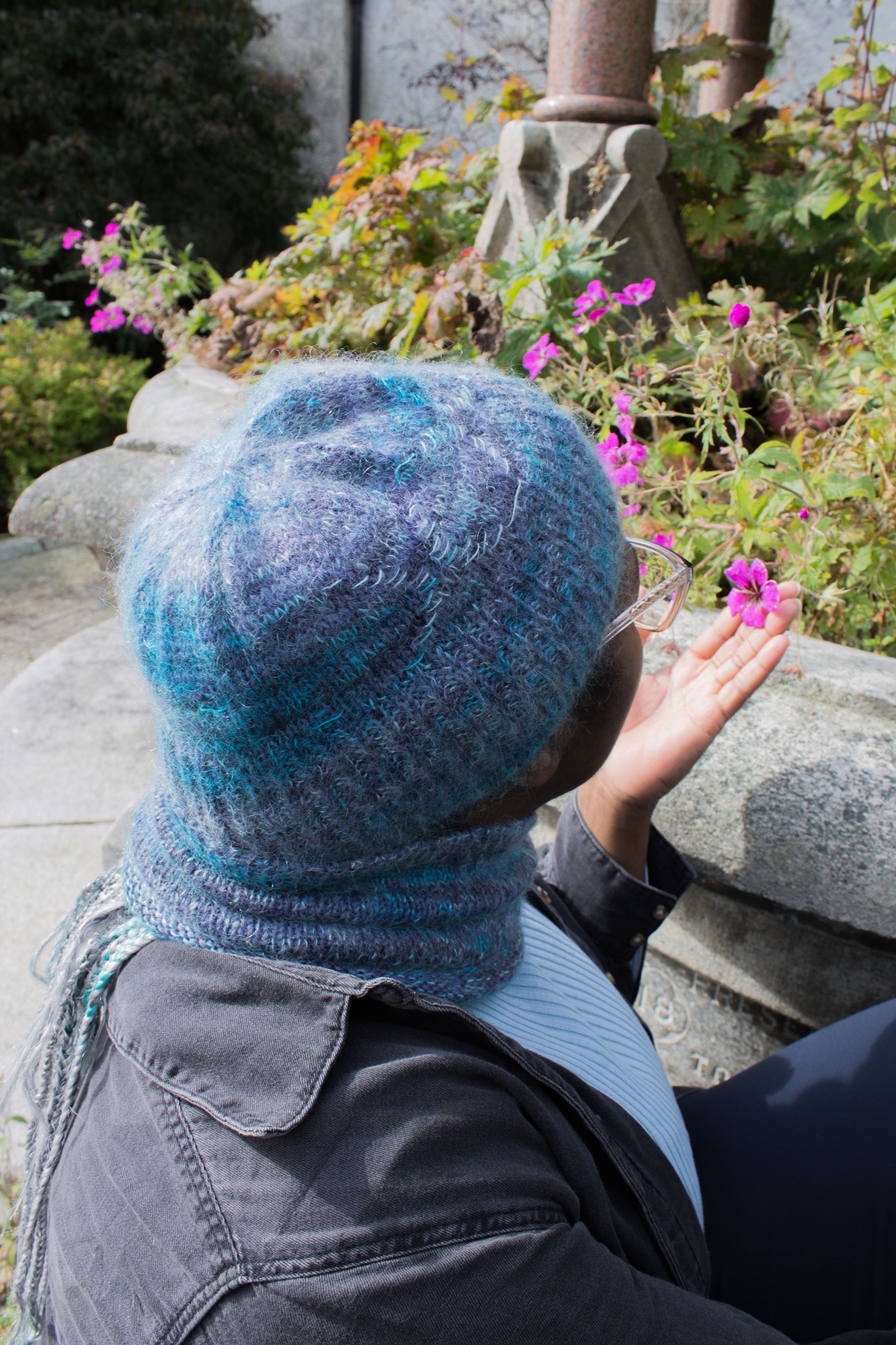 A person gently lifting a flower to their face wearing a hat and cowl in blue with a delicate triple moon pattern on them