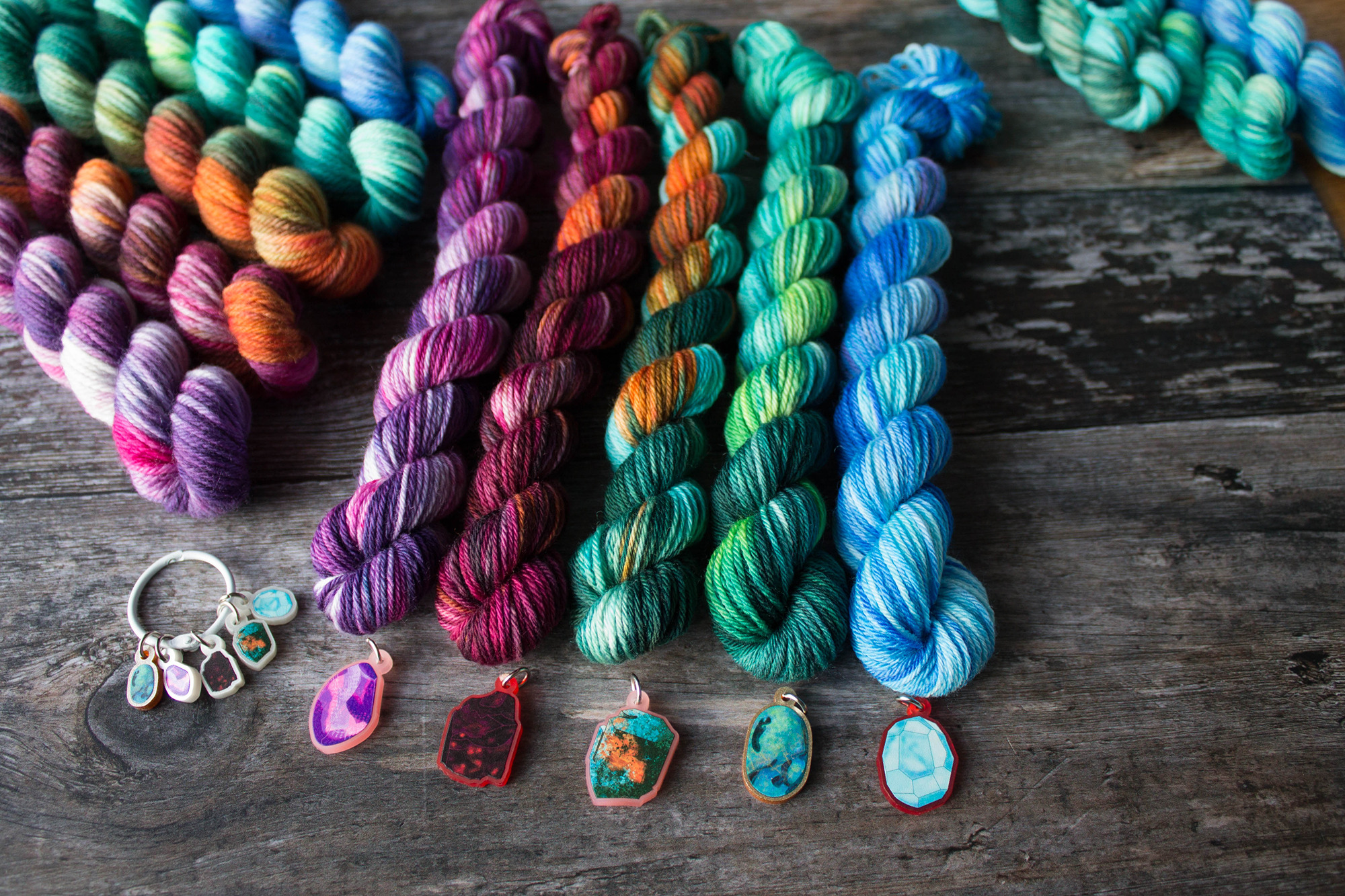 Two sets of mini skeins in 5 different colours with large charms that the colours are drawn from in front of them. to the side is a smaler set of stitchmarkers on a ring. the colours are purples, orange, greens and blue