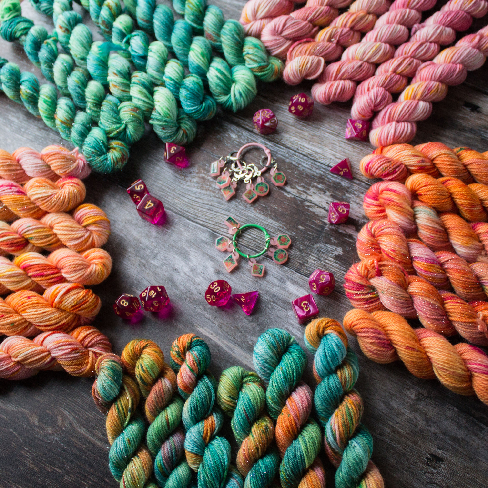 5 different colourways on mini skeins in a pink to green gradient are arranged in sets of six around some pink polyhedral dice and 2 stitchmarker sets that have dice designs in the same colour of the yarn.