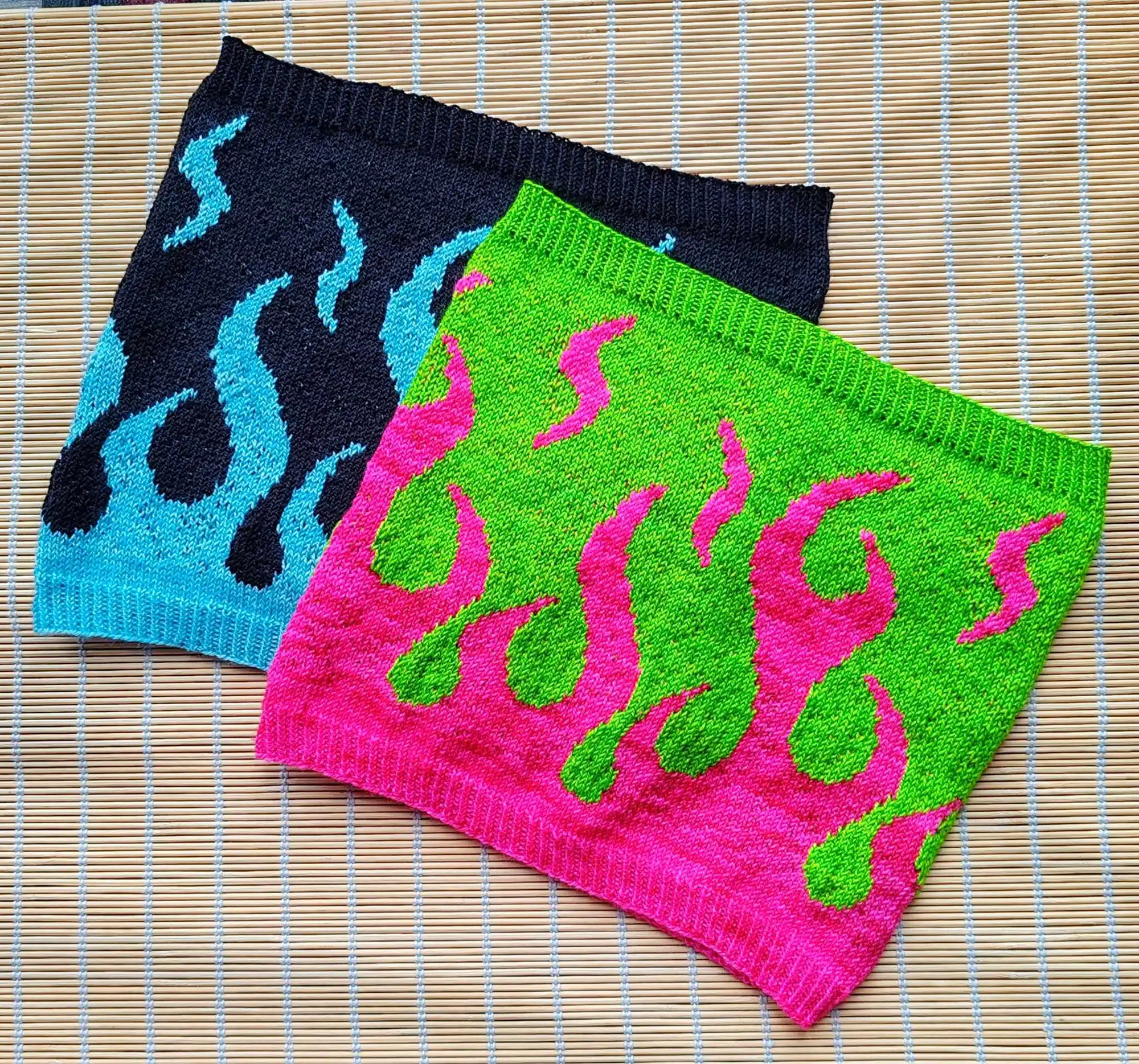 Two cowls on a surface both with the same colourwork design of flames climbing up the cowl. One cowl is in Navy and light blue yarn and the front cowl is in bright pink and Intense green yarn.