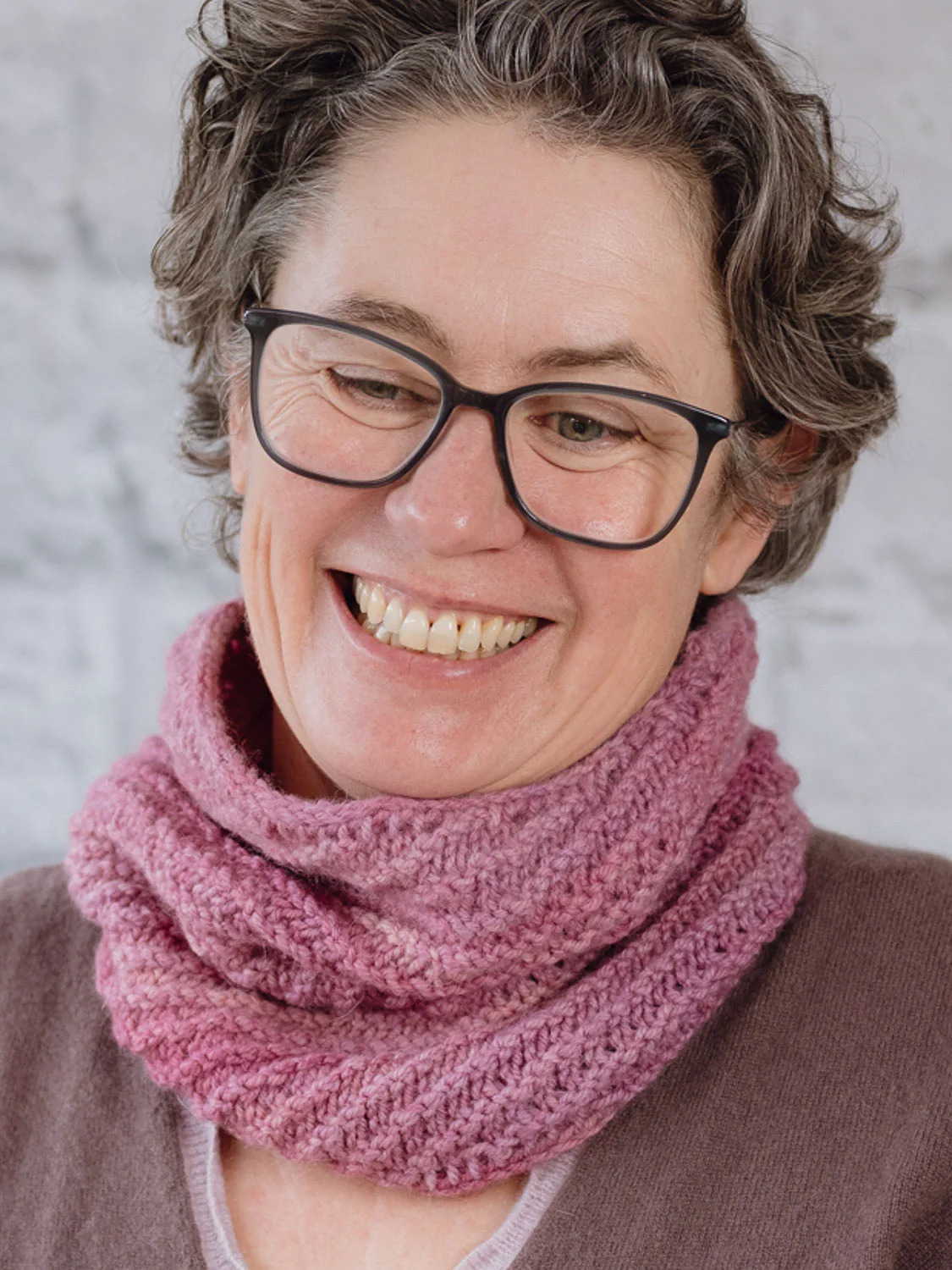 A white person smilling wearing a pink texture knitted cowl