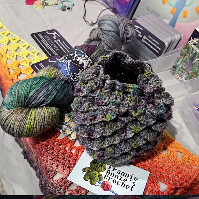 a dragon scale style dice bag crocheted in Fingal's cave featuring grey with pops of lichen green, plum, and bottle green. it is on a business card that reads frannie annie's crochet with a sack puppet and yarn ball logo. next to it is a skein of the yarn and Johnson ofair business cards. they are all on top of a crocheted gradient shawl. 