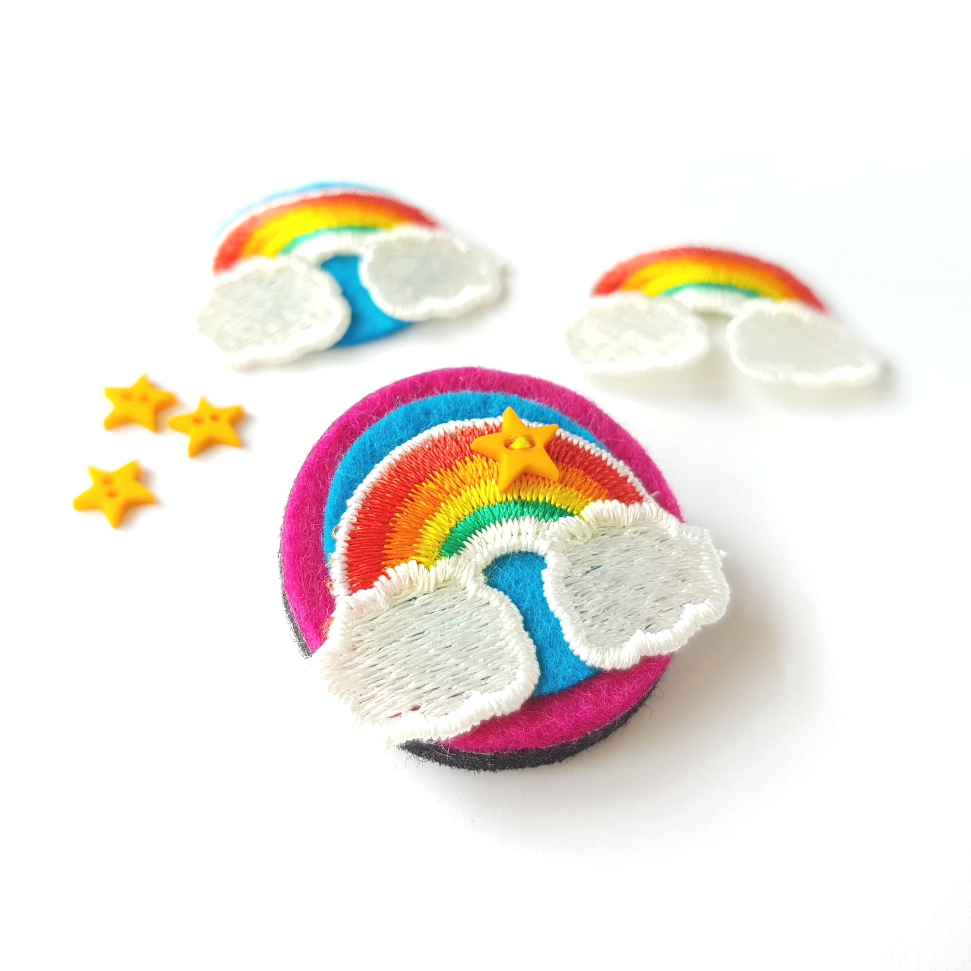 A small, circular, hot pink felt brooch with a stitched rainbow motif and a tiny yellow star shaped button