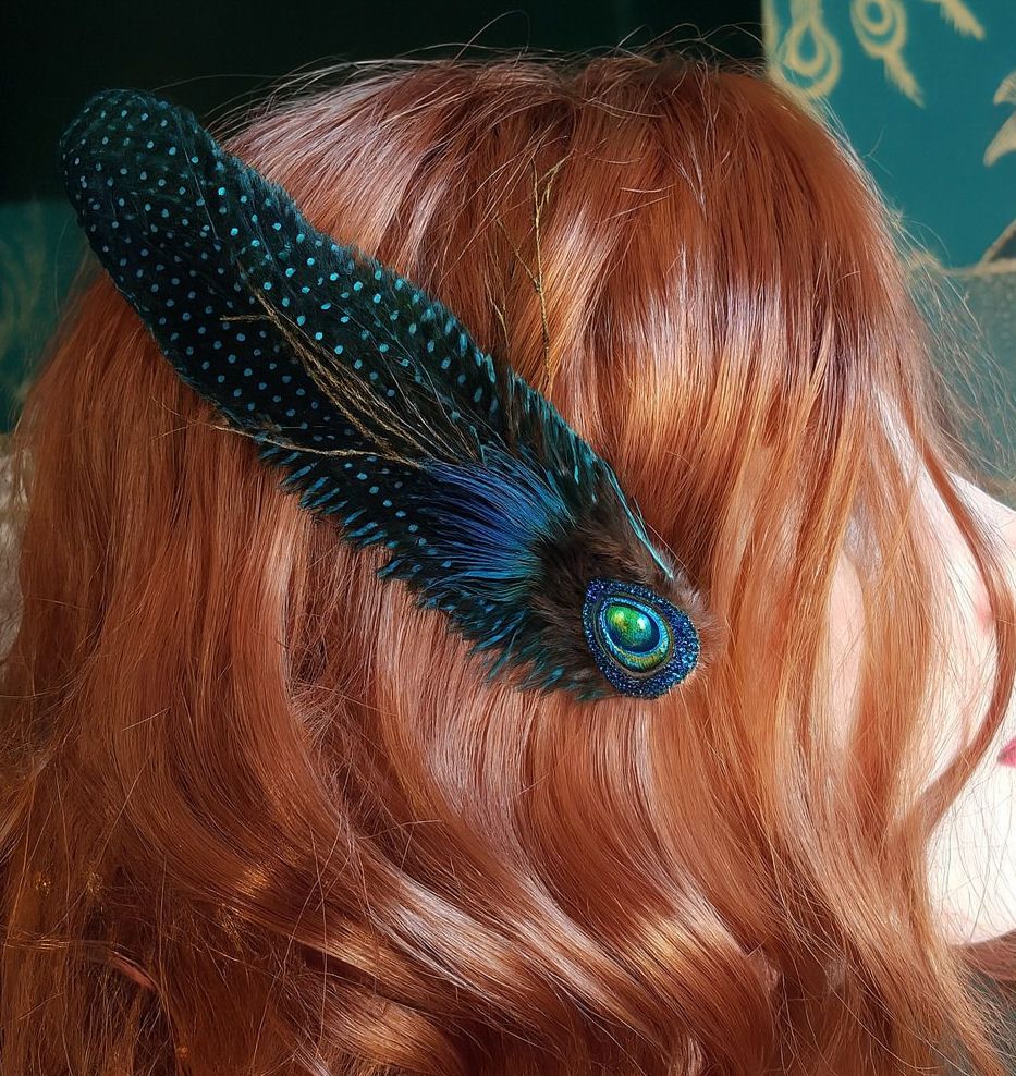 A woman with long, wavy auburn hair wears a vibrant peacock feather hair cl