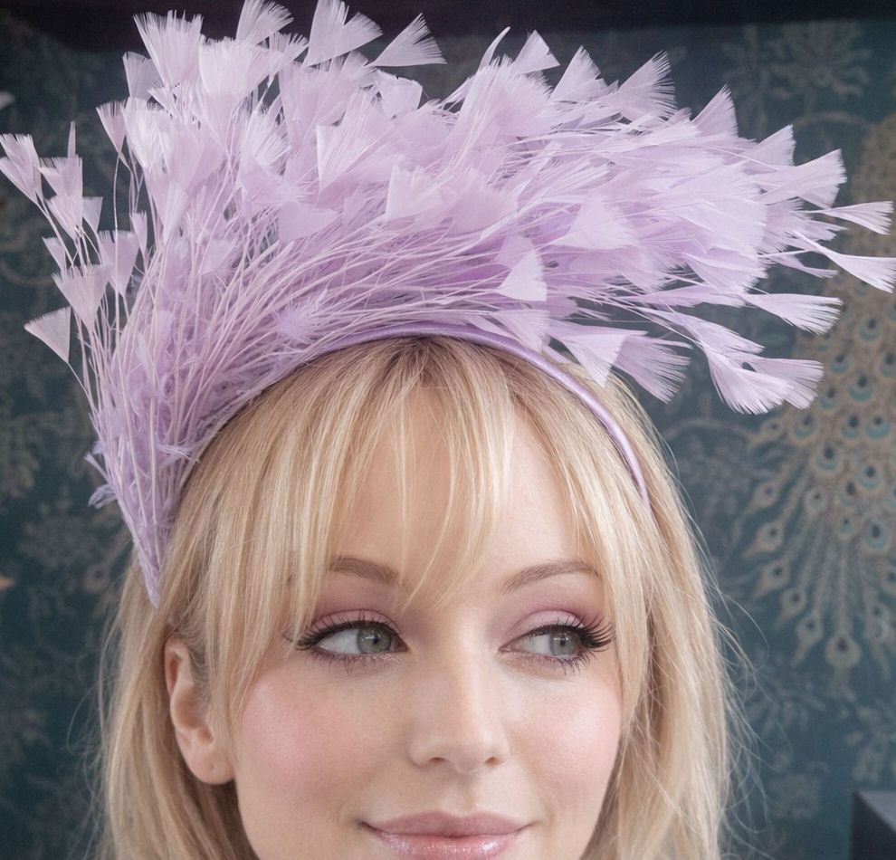 A blonde woman wearing a dramatic lilac feather fascinator looks slightly t