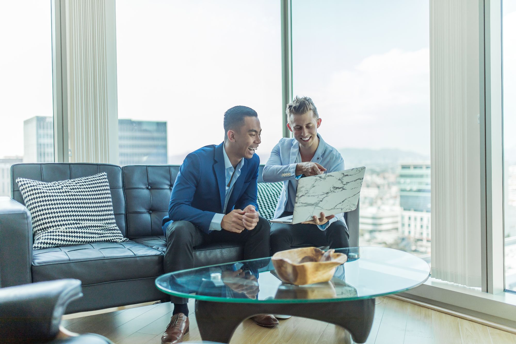 Two men in a highrise apartment smiling, looking at the same laptop screen