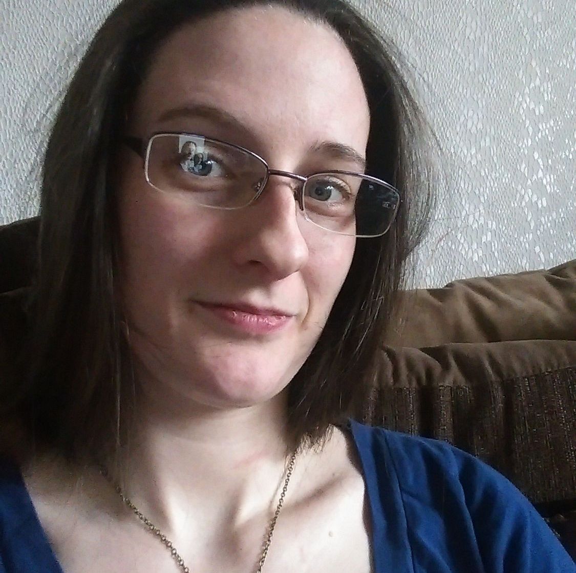 A headshot of Caz. Her hair is loose and dark brown. She wears glasses and a medium blue top.