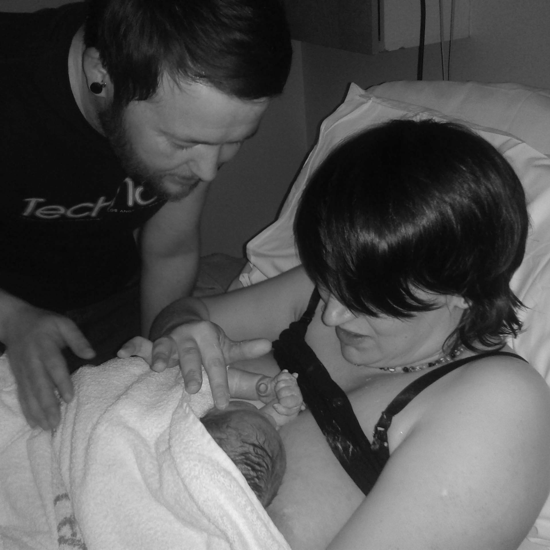 A woman holds her newborn baby to her chest, and touches her face with a finger. Her partner looks down on them both.