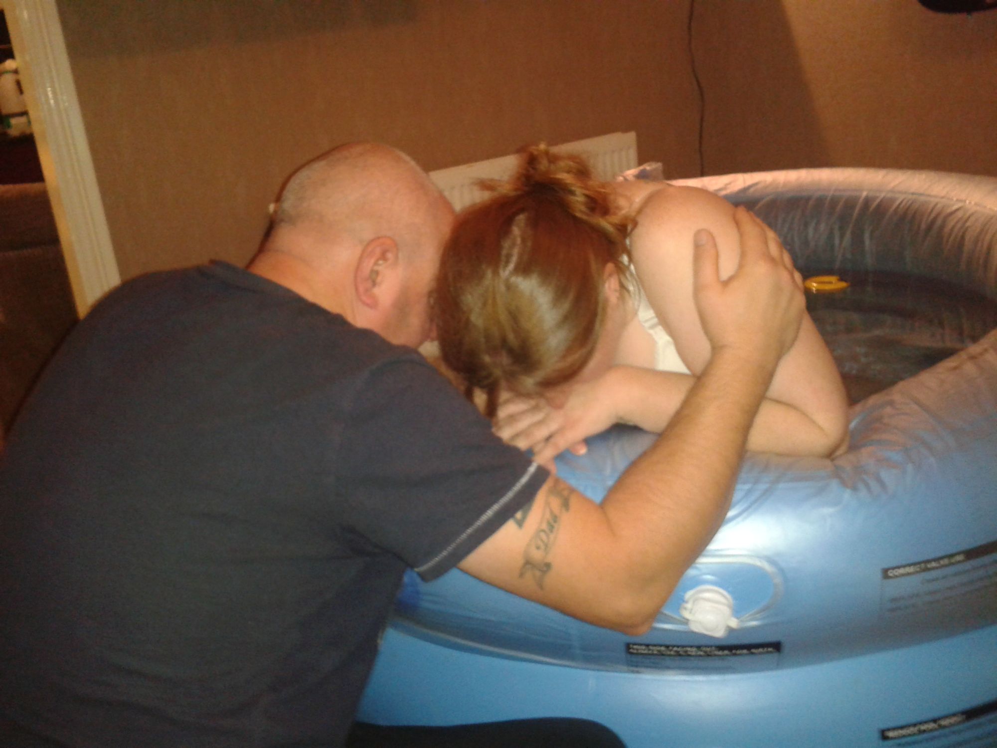 Image of a labouring woman in a pool. She is leaning her elbows on the side and being supported by her partner.