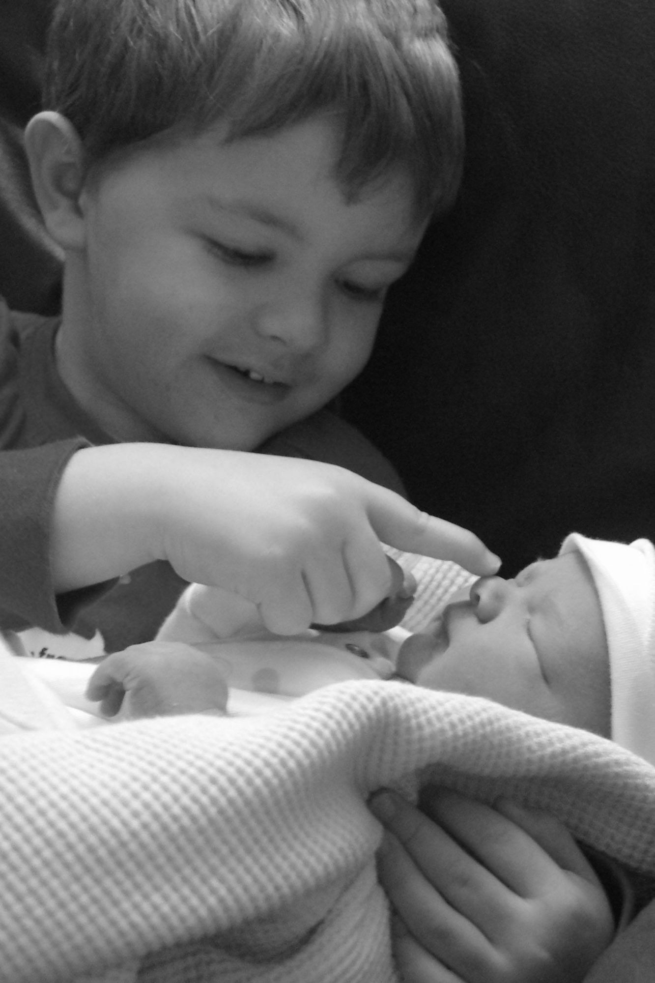 A black and white image of a toddler cuddling a newborn baby wrapped in a blanket. The toddler is touching his brother's nose.
