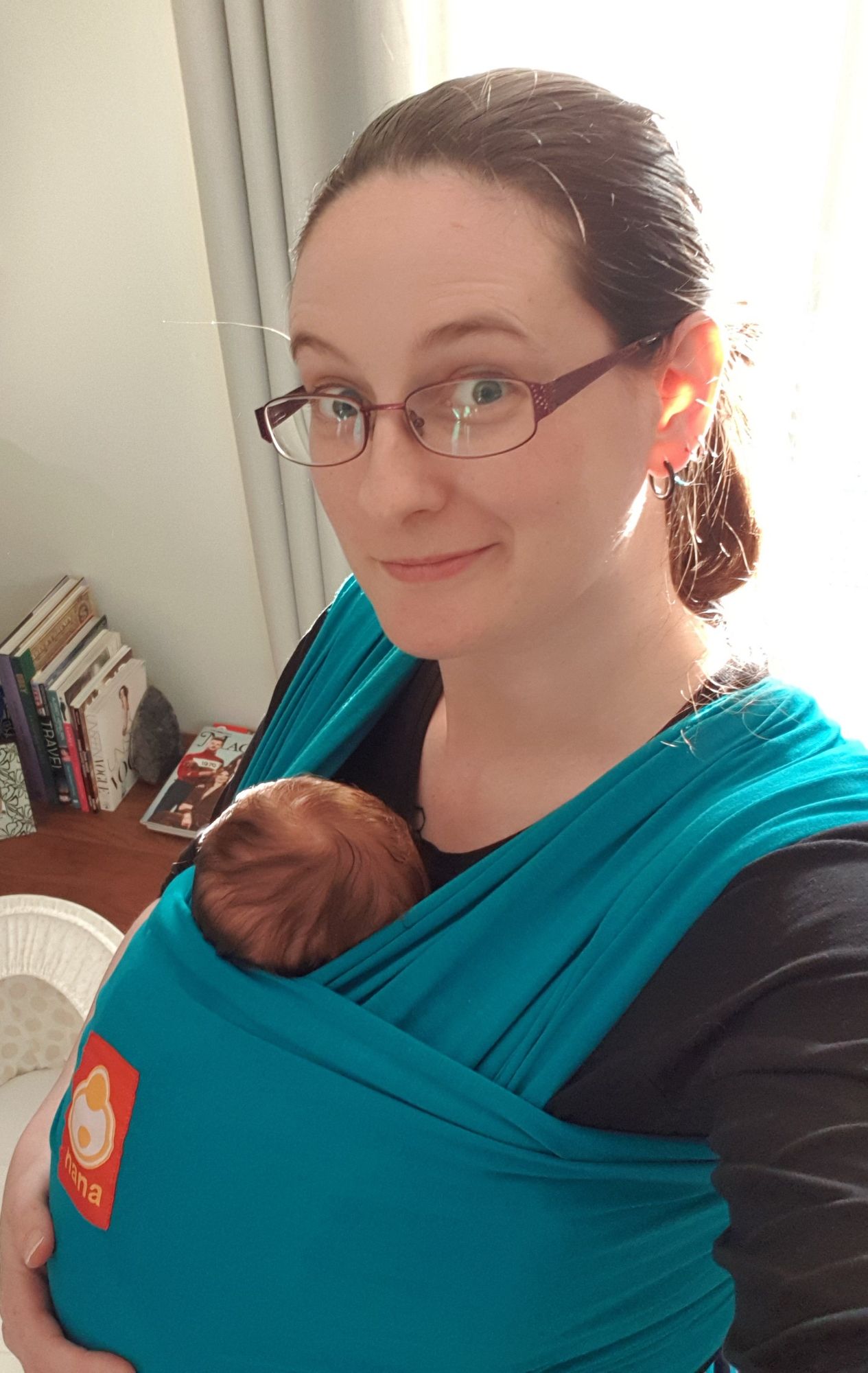 Caz is wearing a newborn baby in a teal stretchy sling. She is smiling at the camera.