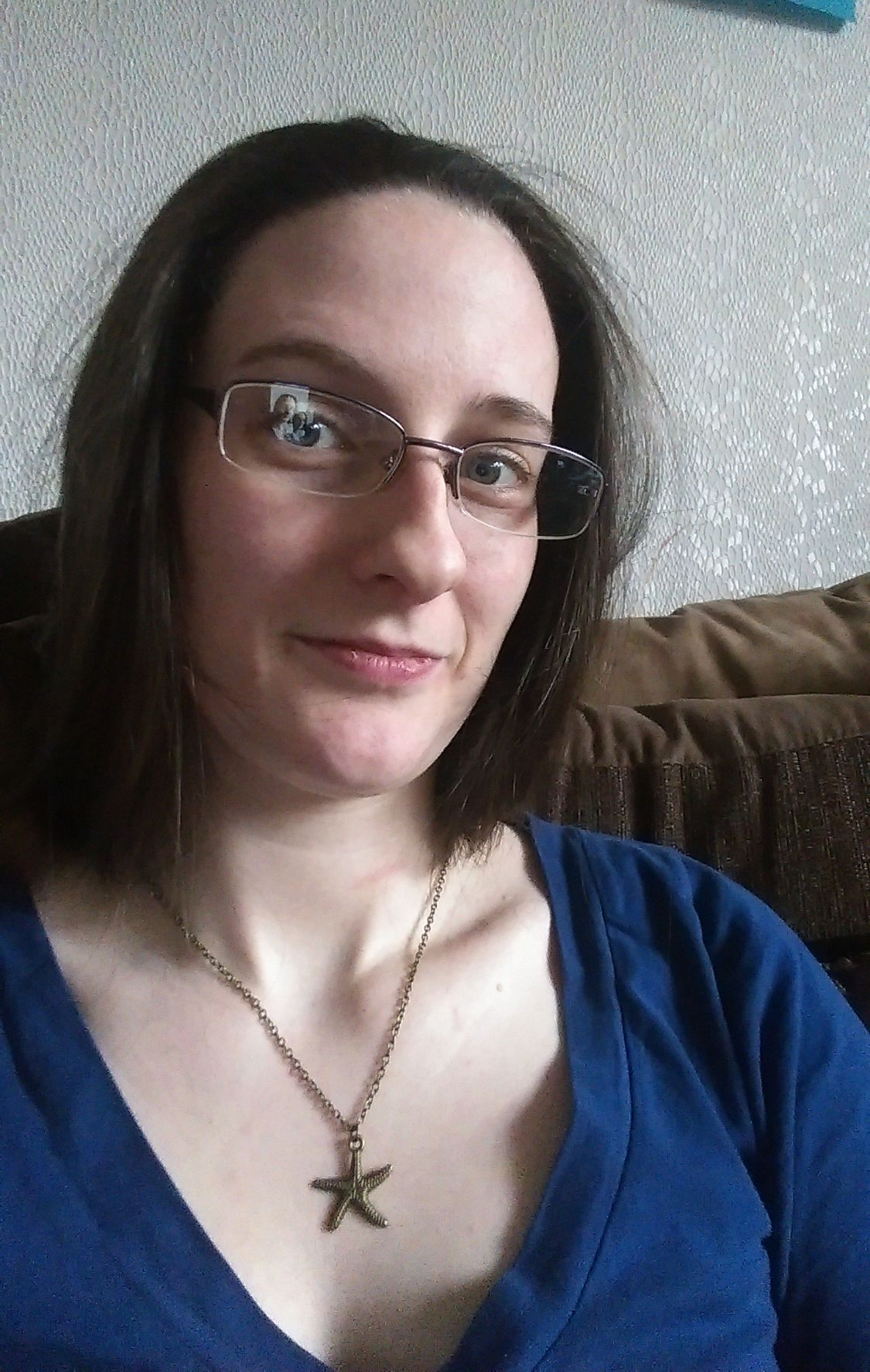 An image of Caz sitting on a sofa. She wears glasses. Her hair is loose and she is wearing a blue top and a necklace with a starfish on it.