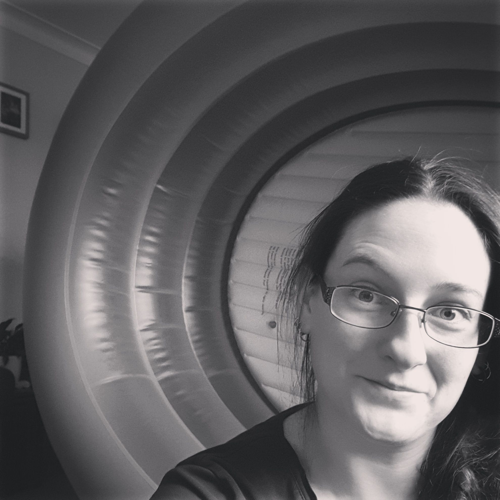 A black and white image of Caz looking into the camera. Her hair is plaited forward over her shoulder. Behind her is an empty birth pool on its side.