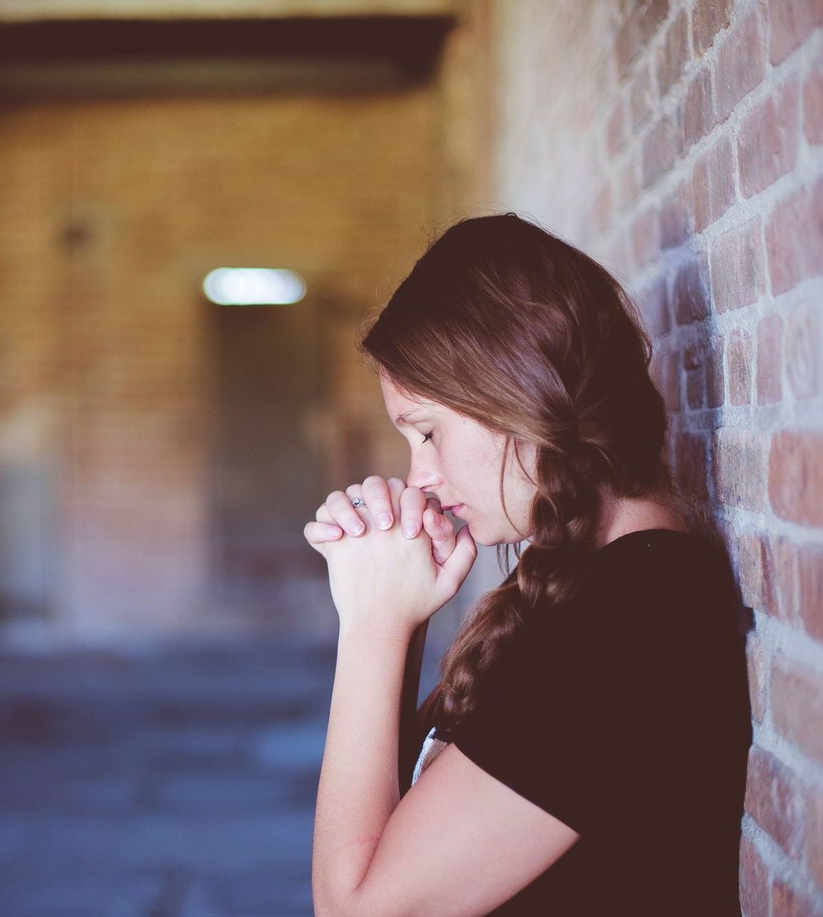 A brown haired woman leans back against a wall. Her eyes are closed and her head bend over her clasped hands as she contemplates something.