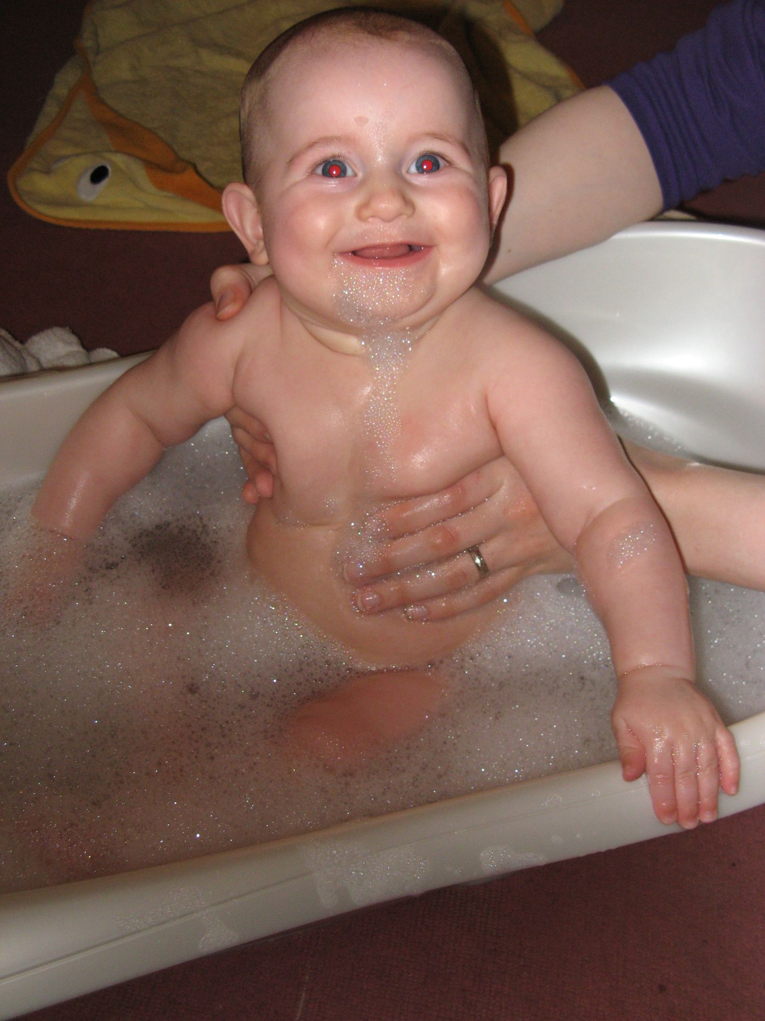 Image of a baby, aged a few months old, grinning at the camera. They are being supported sitting in a baby bath.