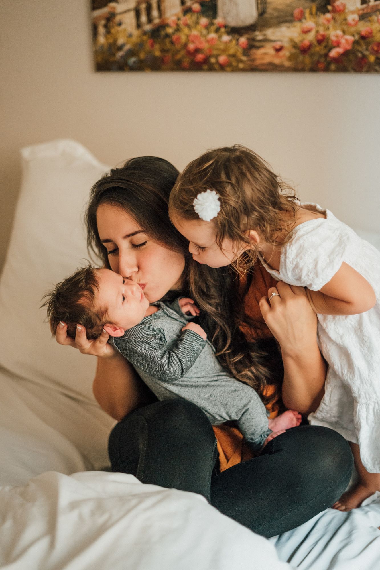 Image of a mother kissing a new baby. A toddler is leaning over her shoulder to kiss the baby too.