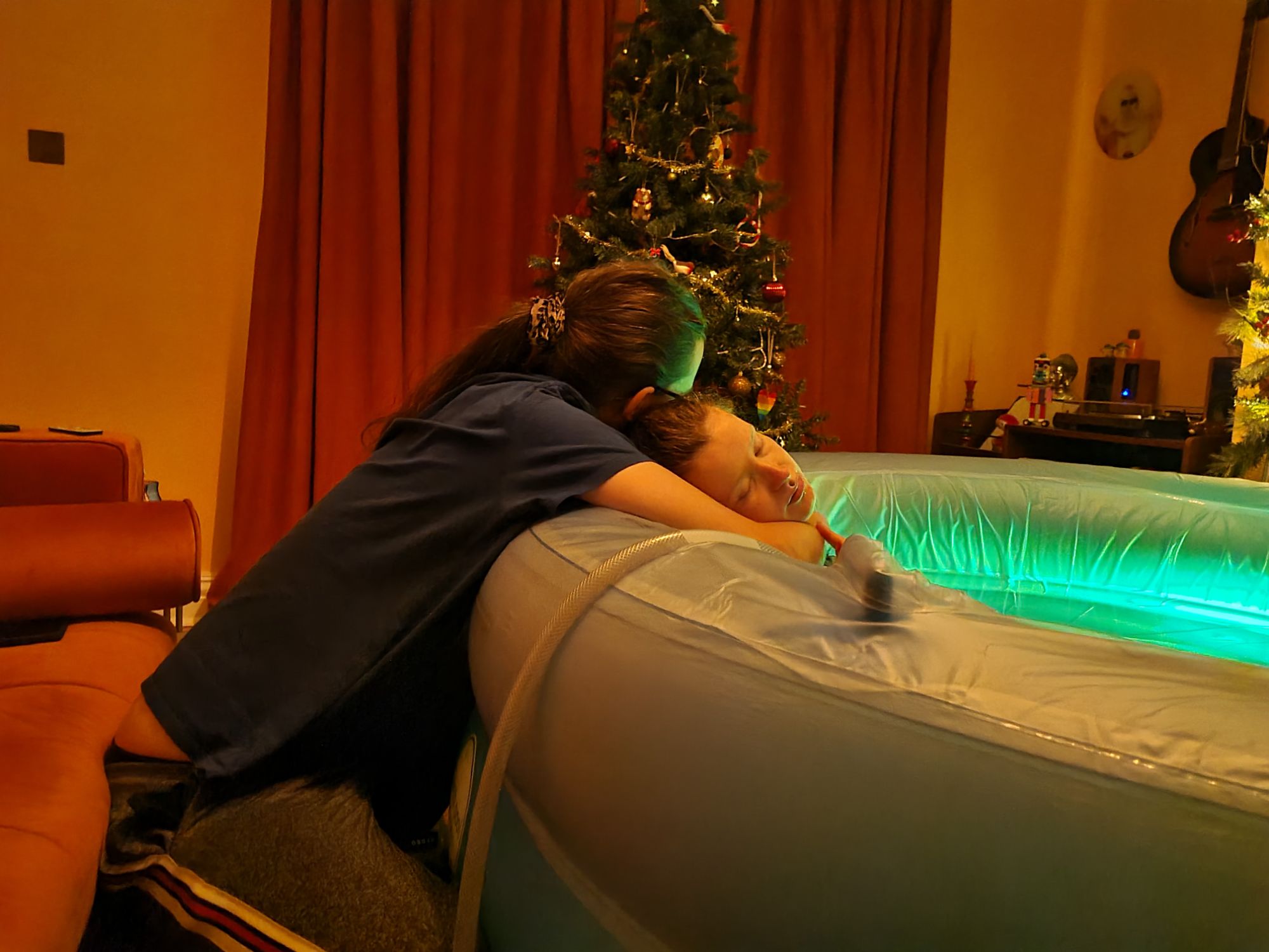 Image of a woman reclining back in a pool, with her partner hugging her from behind over the edge of the pool. There is a Christmas tree in the background.