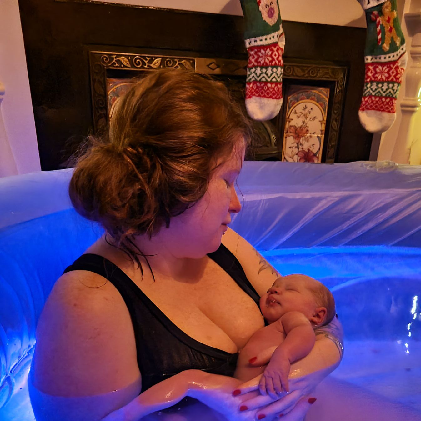 A woman sitting in a birth pool holding a newborn baby. The water is illuminated blue from the submersible light in the water.