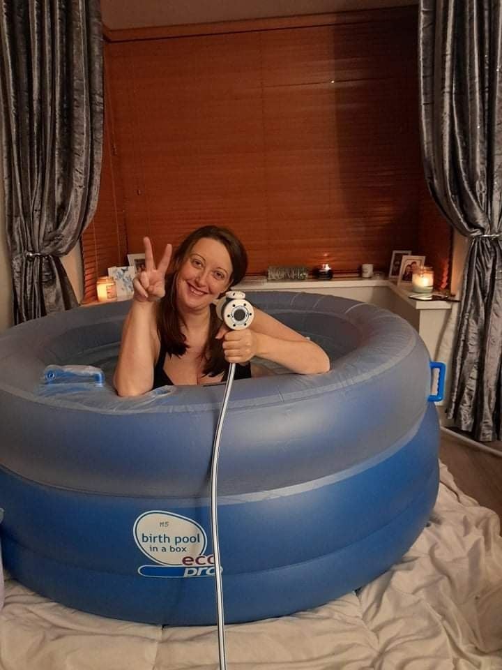 A mini pool in a client's living room. A woman is sitting inside, wearing a black bra. She is holding gas & air in one hand and making the peace sign with the other, and grinning at the camera.