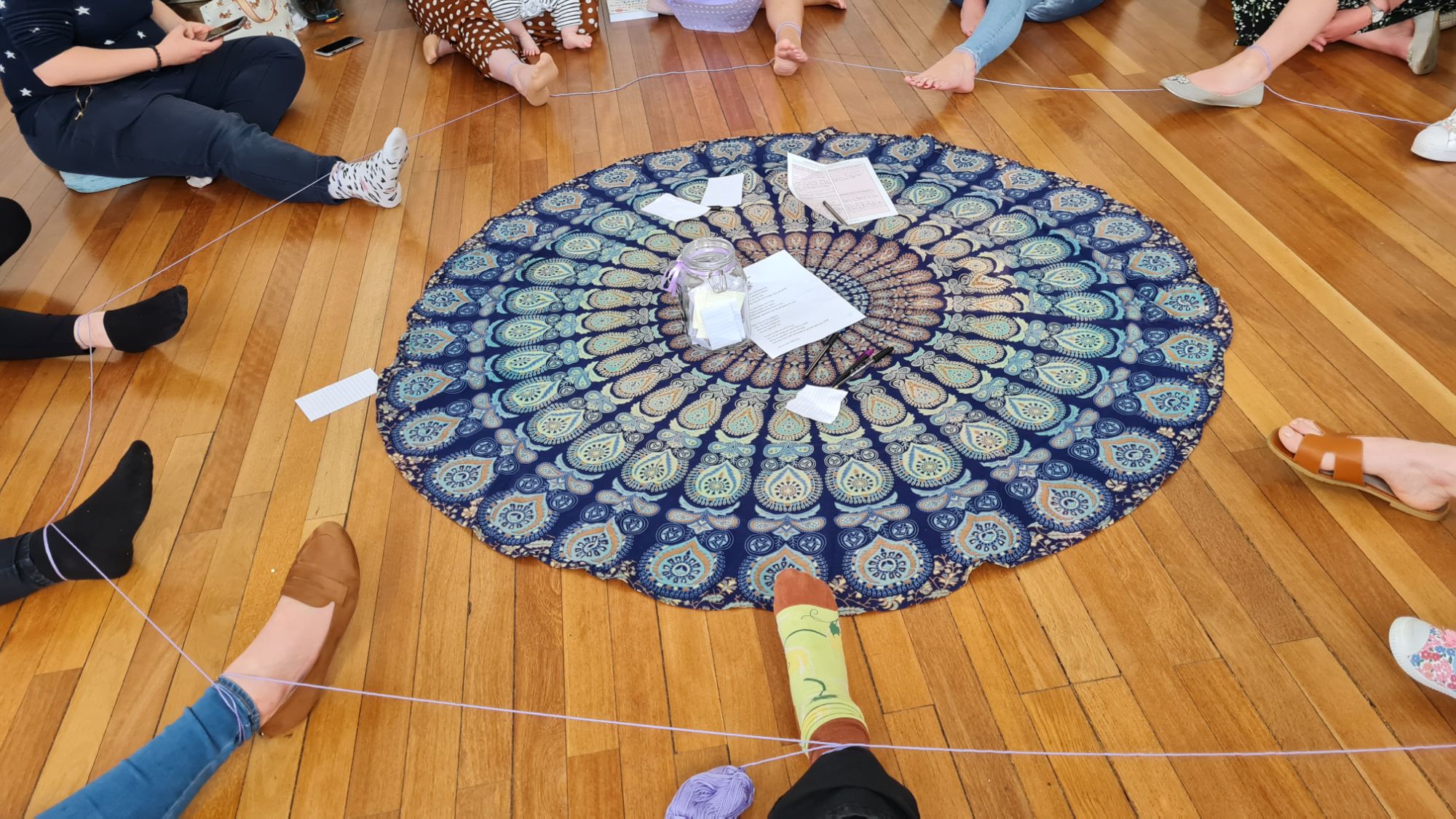 A circular blanket is in the centre of the photo. Around it there are multiple people sat on the floor with one leg extended into the centre. Their ankles are loosely tied together with yarn.