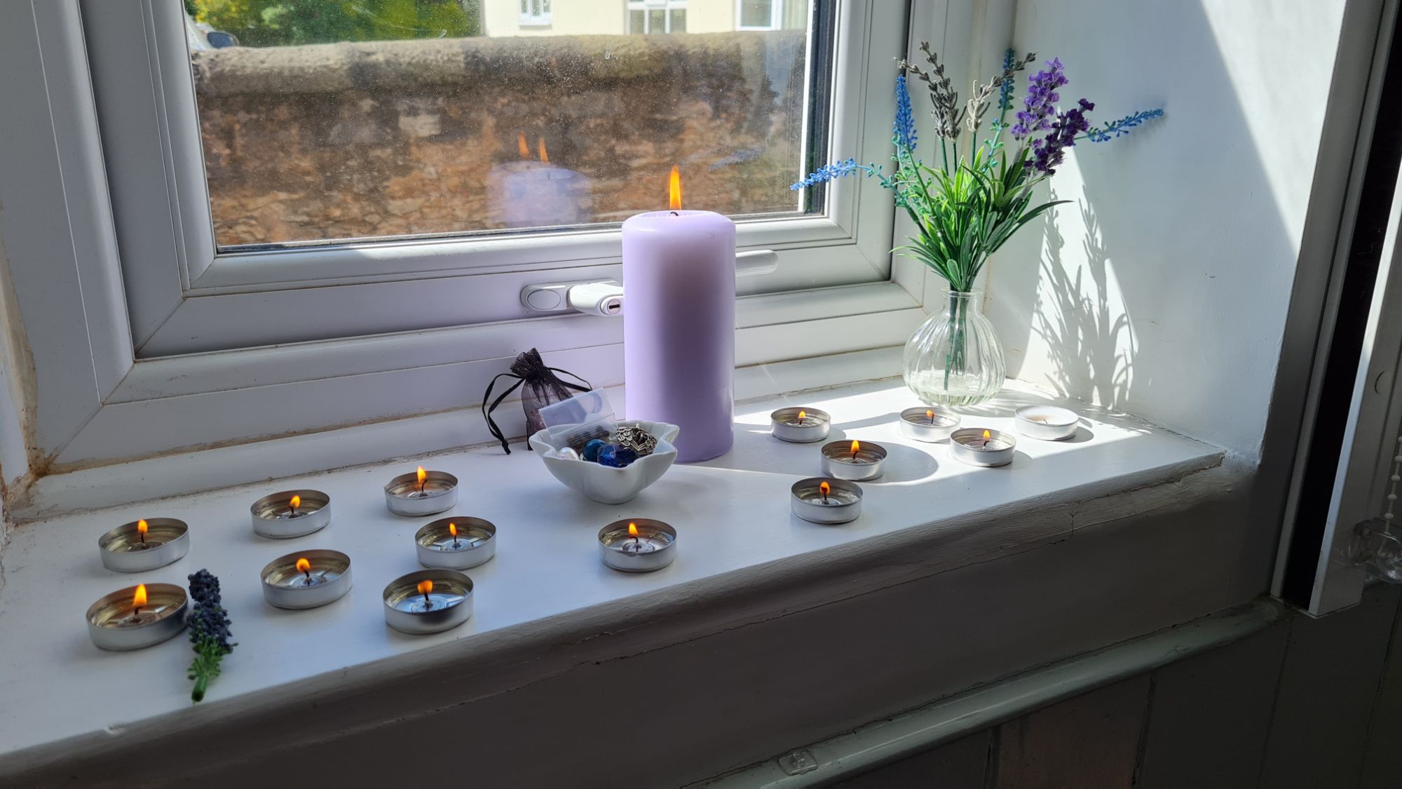 A lit lilac pillar candle on a window ledge, surrounded by lit tealights. There is a small vase of flowers in the corner of the window ledge.