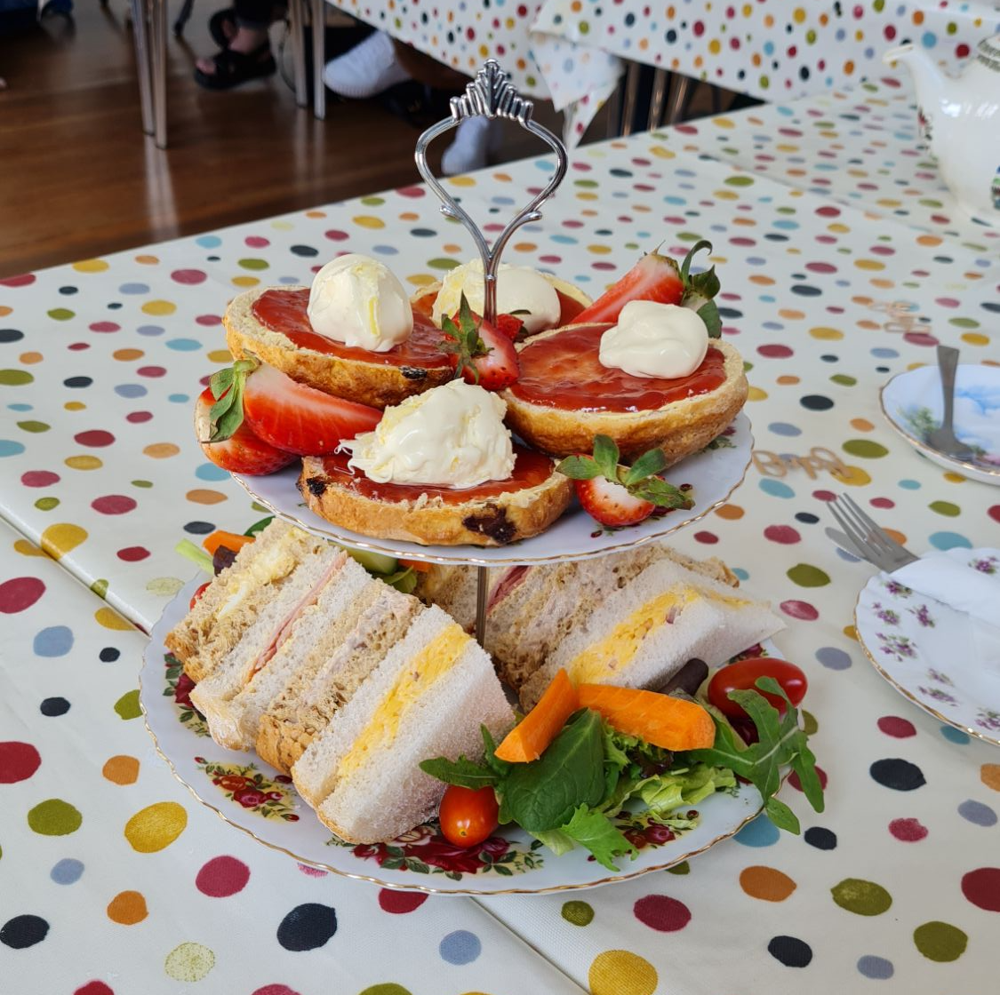 A tiered cupcake stand on a table with a spotted tablecloth. The top tier has jam and cream scones and strawberries on it, and the bottom tier has various sandwiches and salad on it.